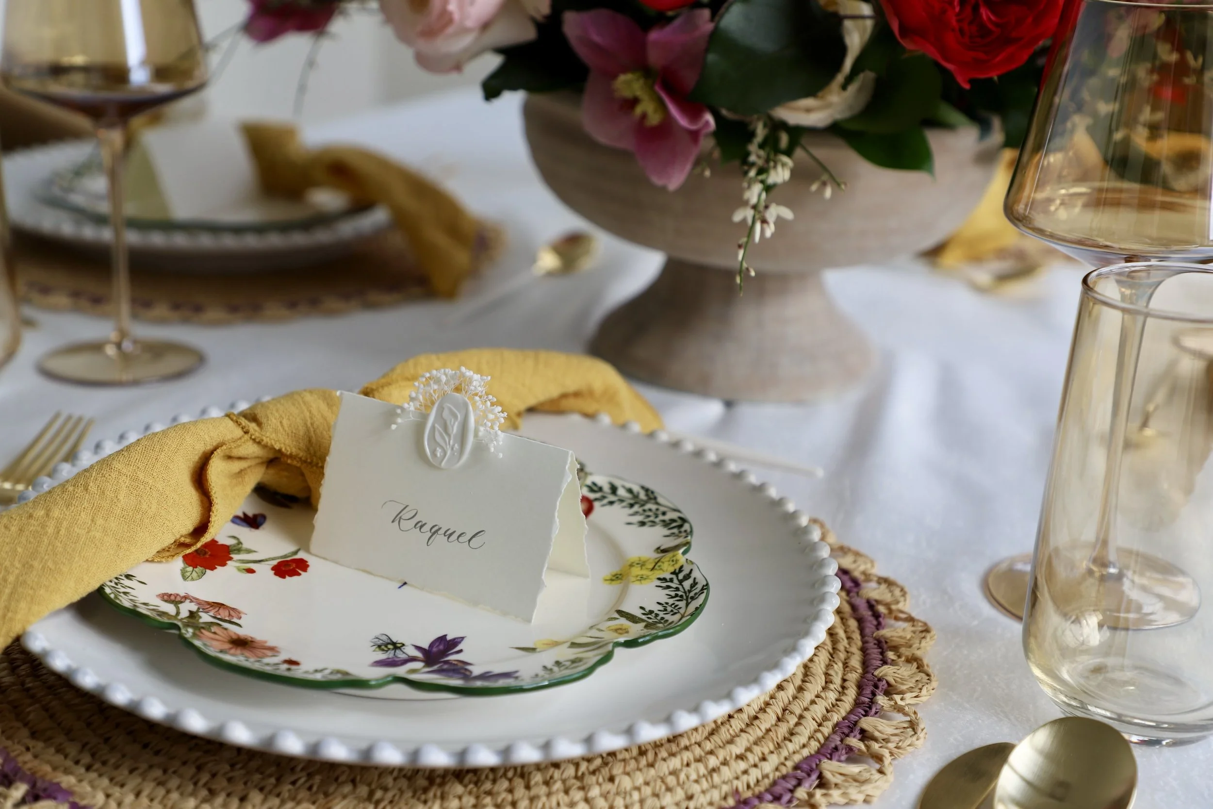 Elegant table setting with a floral arrangement, a decorated plate featuring a name card, gold utensils, and wine glasses, on a white tablecloth.