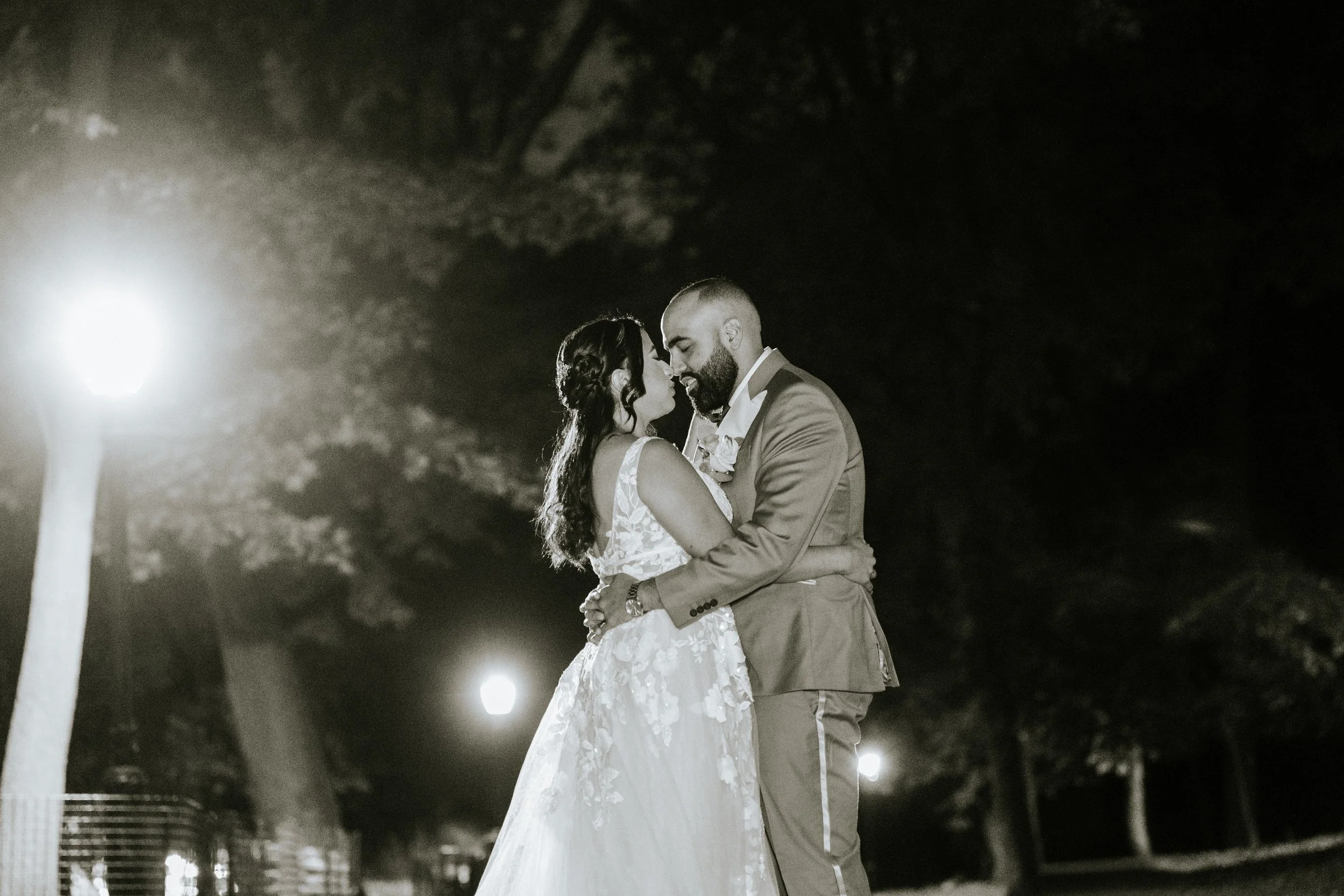 A black and white photo of a newlywed couple dancing outdoors at night, illuminated by streetlights, with trees in the background.