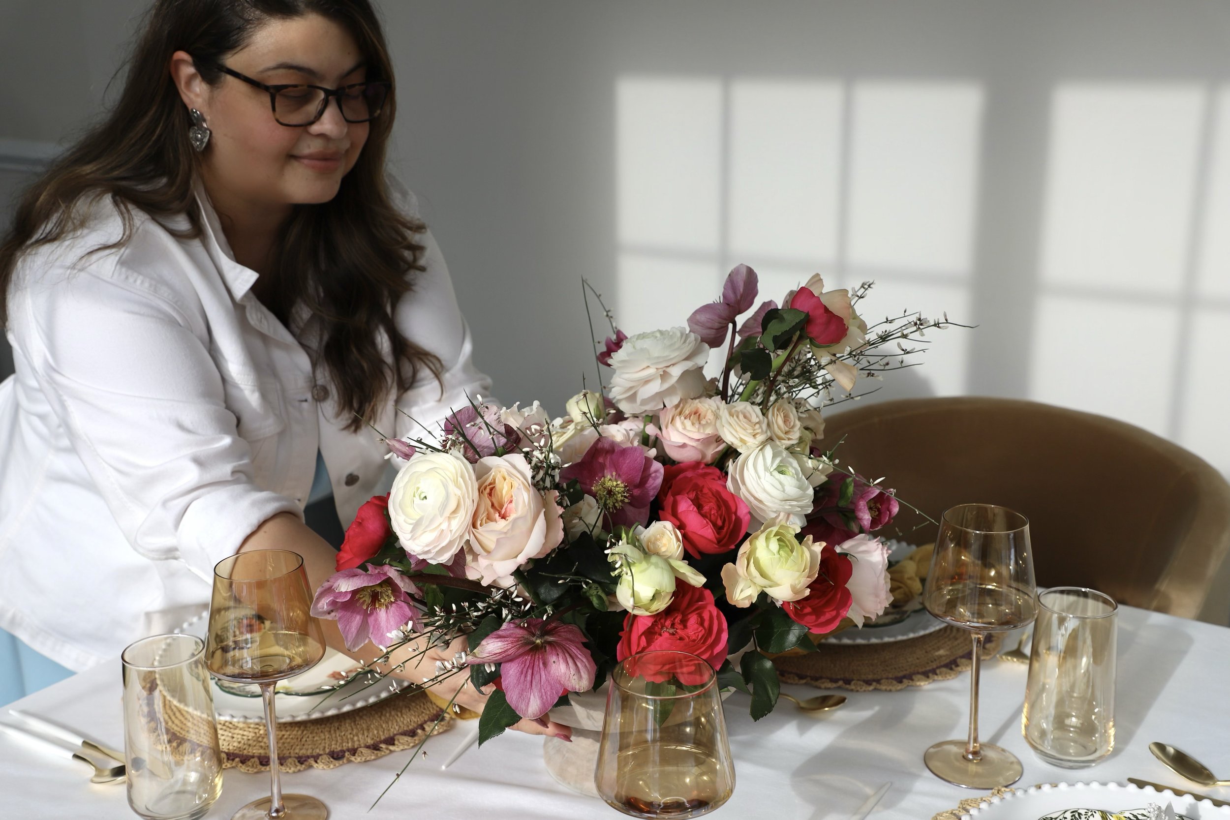 A woman arranging a colorful flower centerpiece on a dining table decorated with gold-rimmed glassware and plates in a well-lit room.