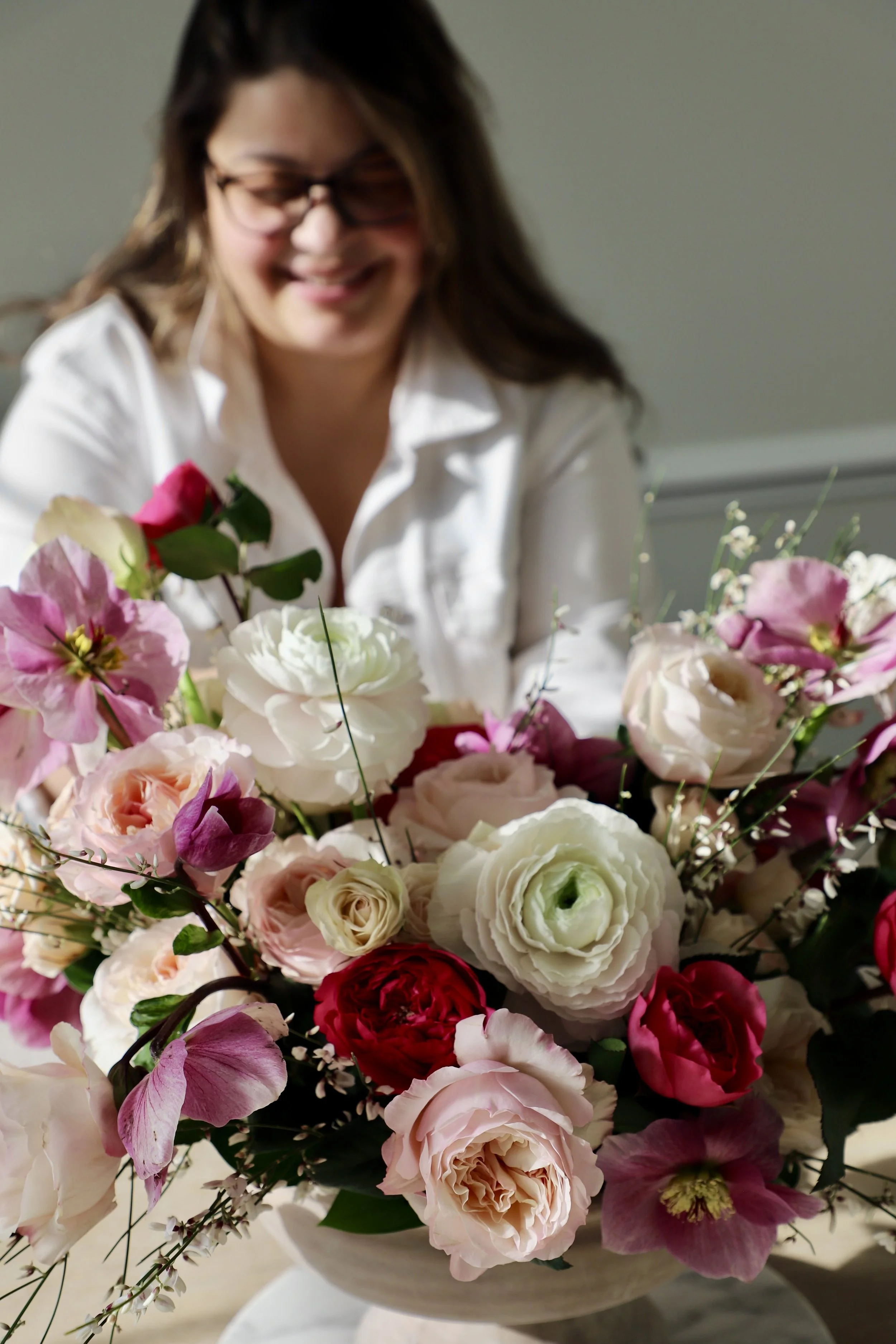 Woman in white shirt smiling at a large bouquet of mixed pink, white, and red flowers in a white vase.