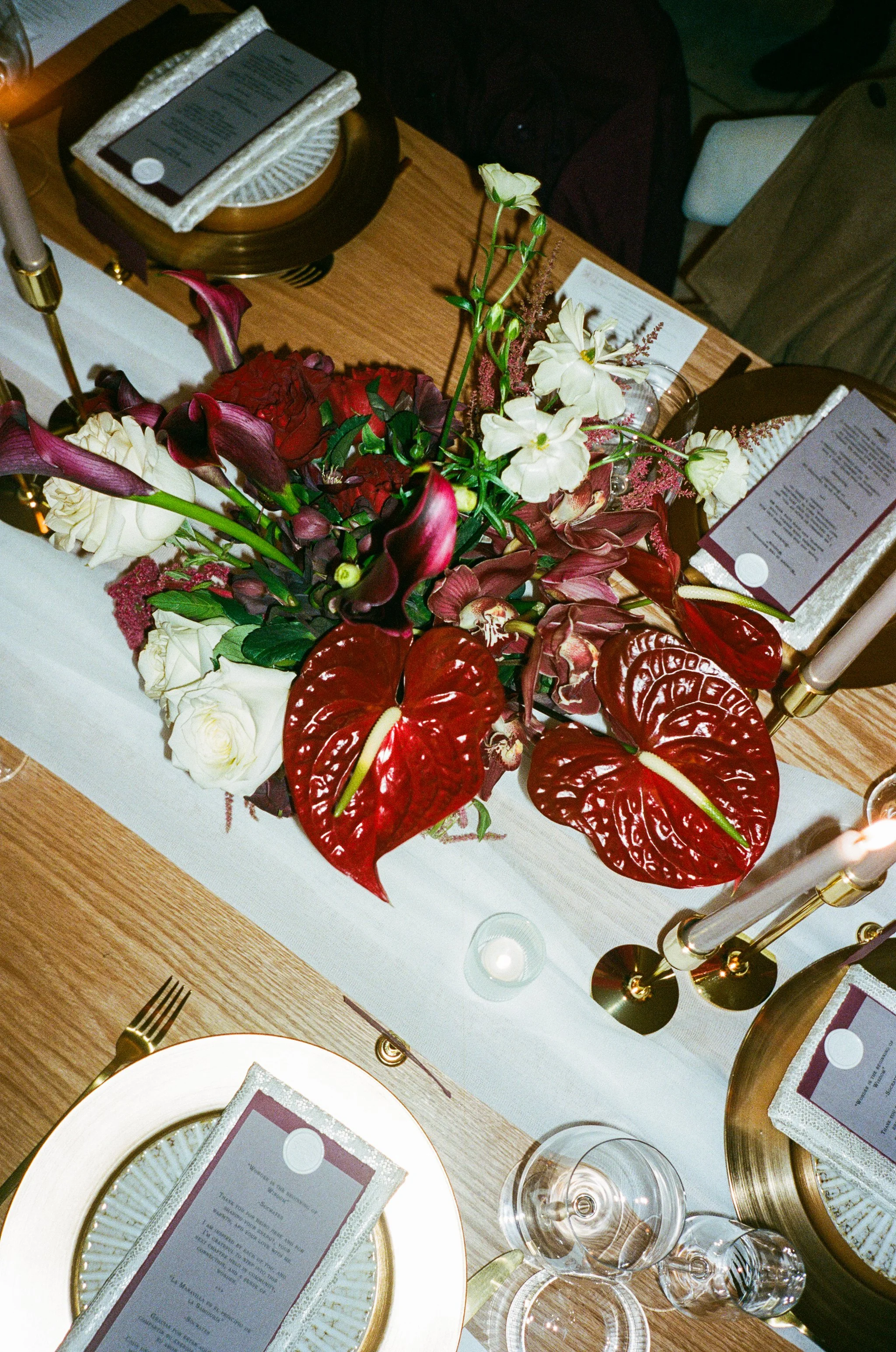 A floral centerpiece with red, white, and purple flowers on a wooden dining table set for a formal event, including white and gold plates, glassware, cutlery, candles, and printed menus.
