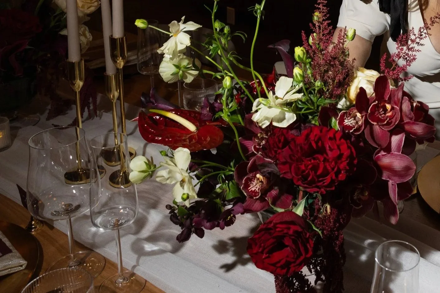 A dining table centerpiece with red, white, and purple flowers including anthuriums, roses, and orchids, accented by tall white candles in gold holders, and surrounded by empty wine glasses. A person wearing a white top with black accents is partially visible to the right.