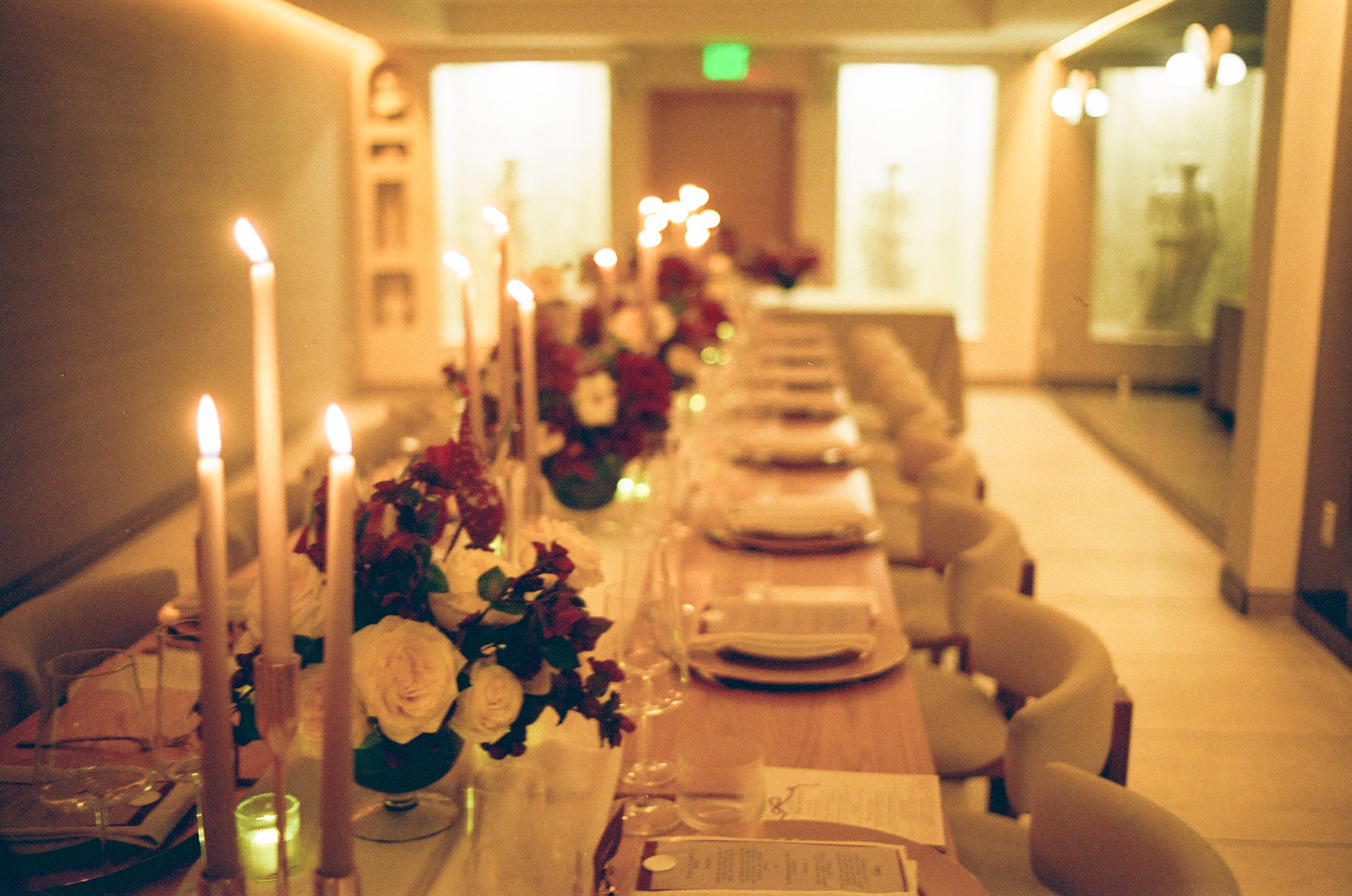 A long dining table set for a meal, decorated with candles and floral arrangements, in a softly lit room with chairs neatly arranged around the table.