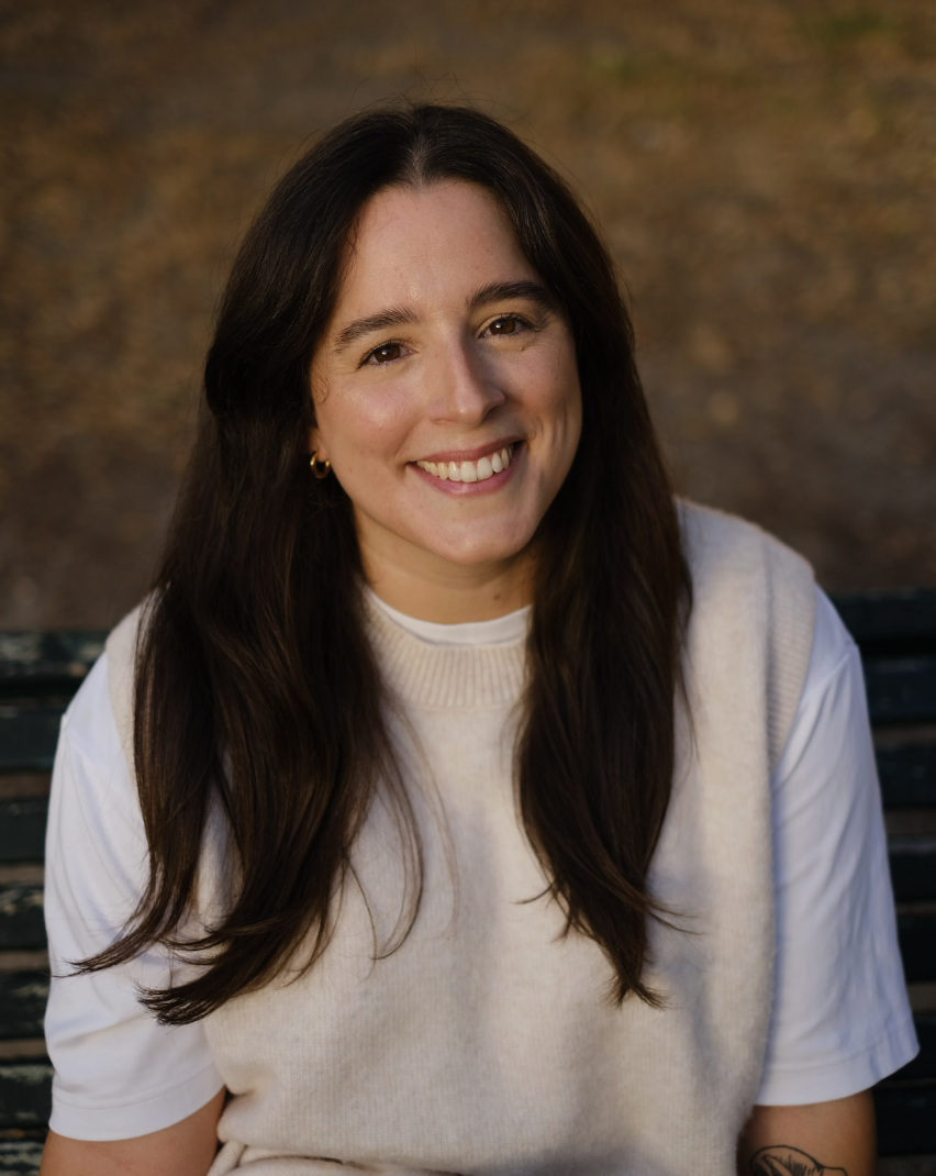 A young woman with long dark hair smiling, sitting on a park bench with autumn foliage in the background.
