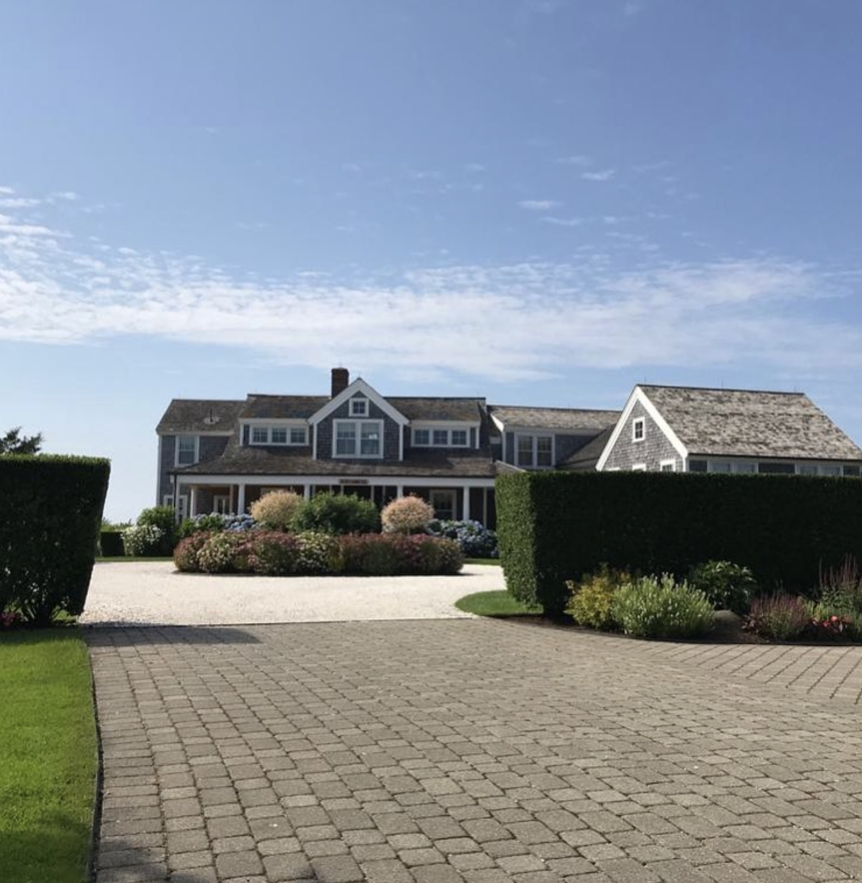 A large house with a garden and driveway, surrounded by well-maintained hedges and colorful bushes, under a partly cloudy sky.