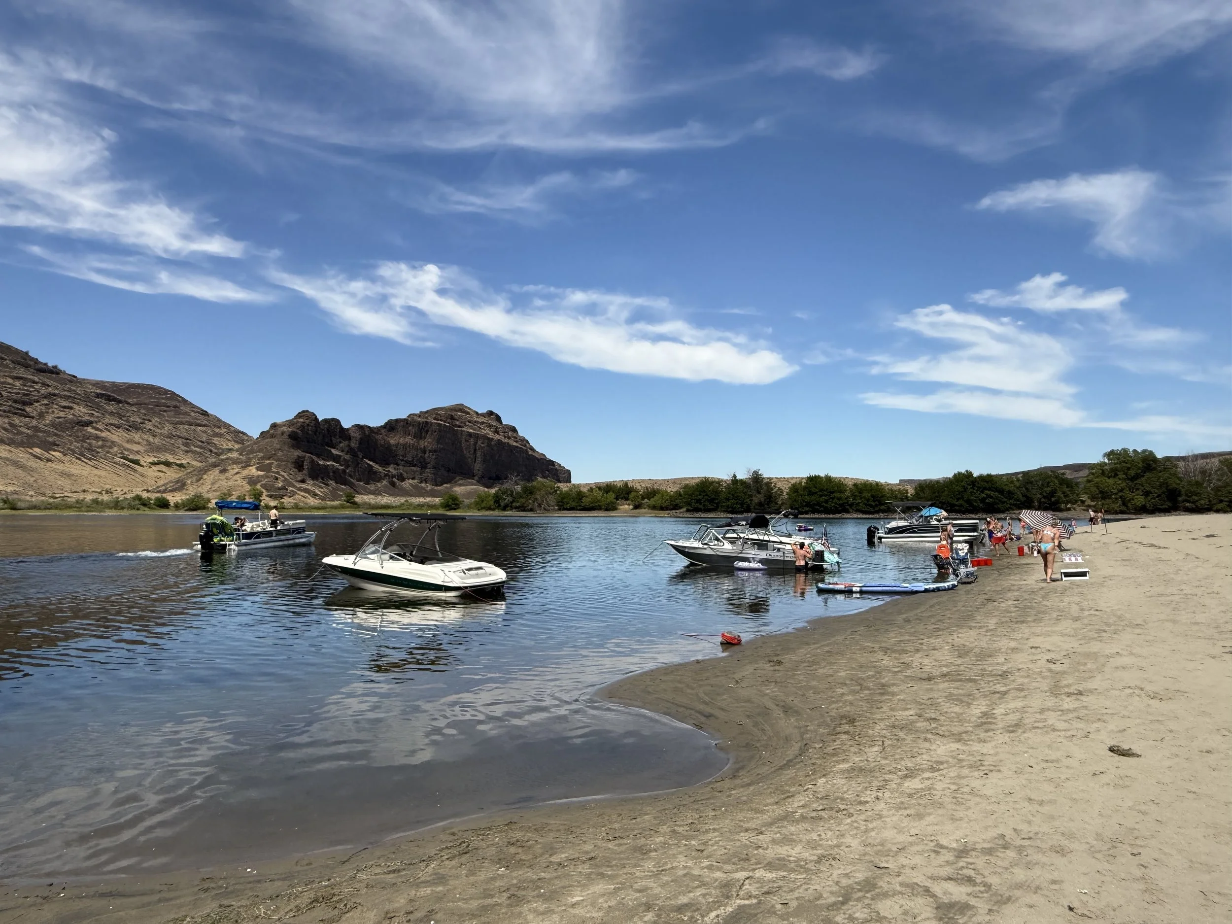 Beach scene with boats anchored near shore, people relaxing under umbrellas, and a rocky hillside in the background under a partly cloudy sky.
