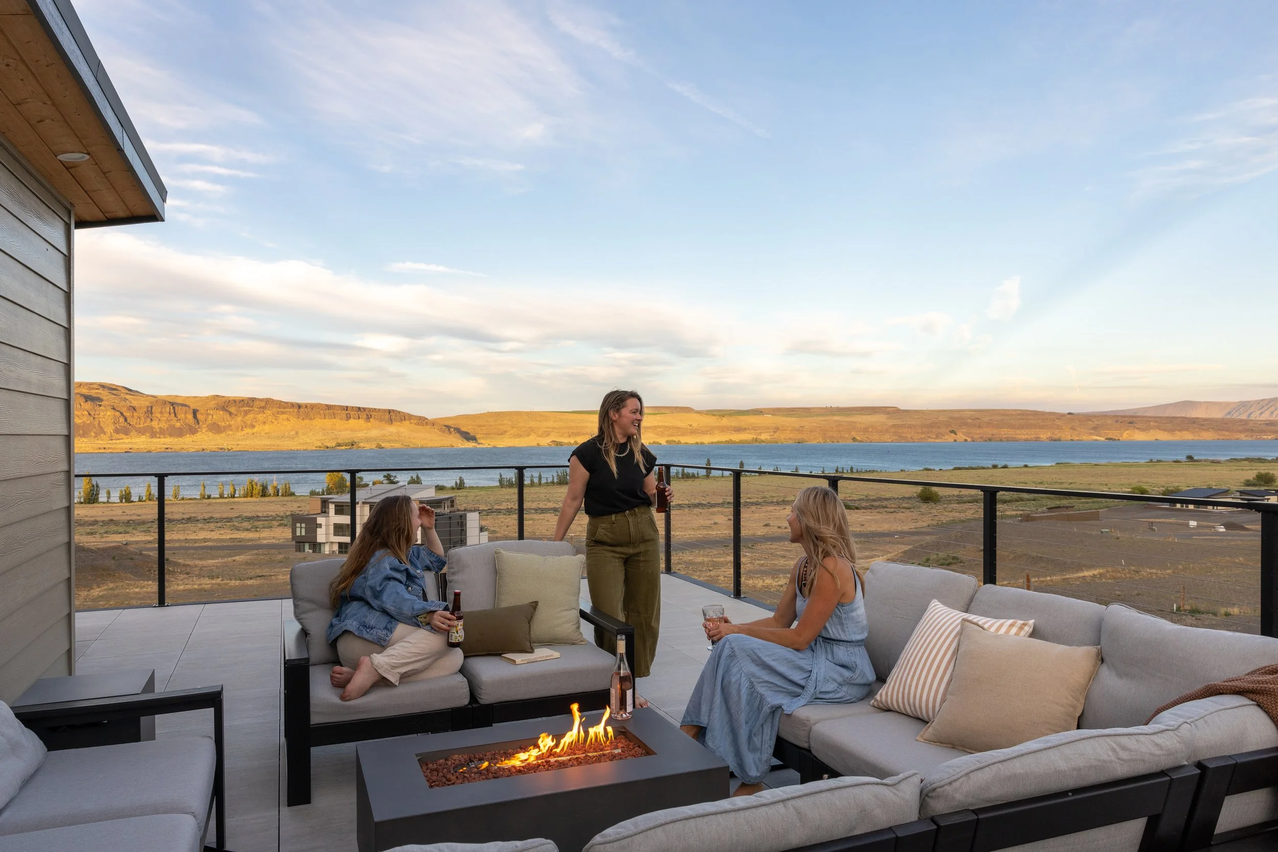 Three women sitting on outdoor sofas, laughing and holding beverages, enjoying a drink next to a firepit, with a scenic landscape of a river and distant hills in the background.