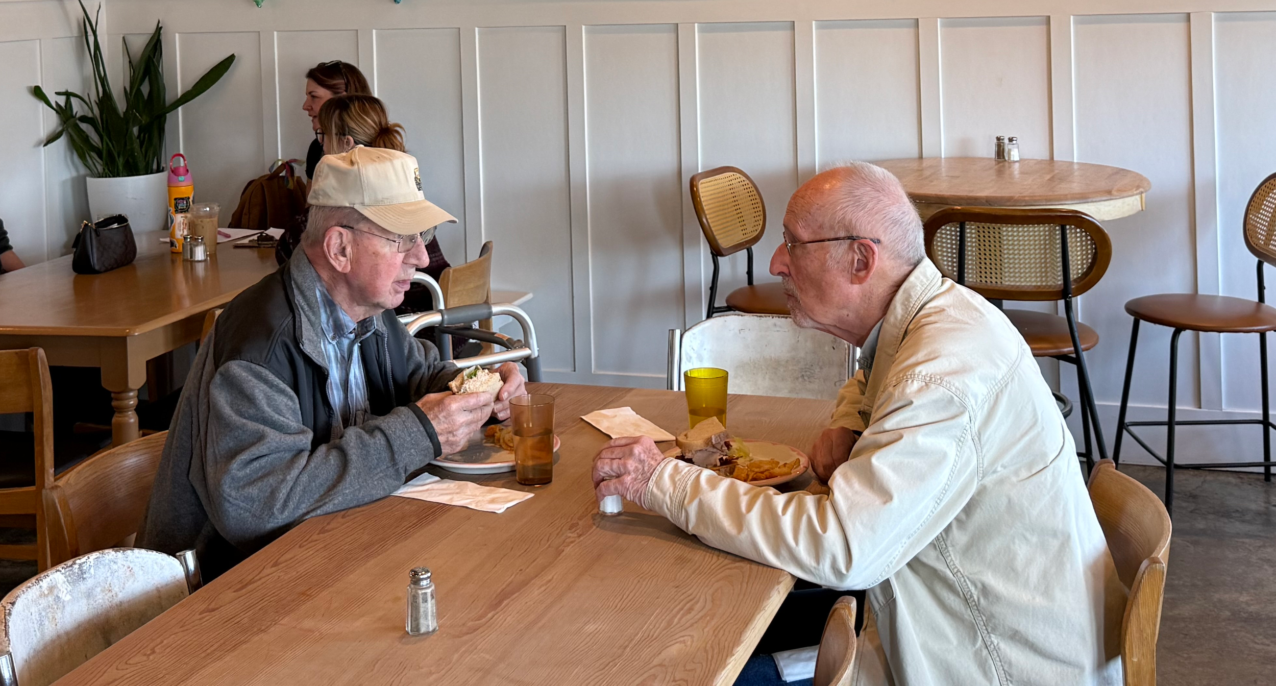 Two older men talking and eating sandwiches at a table with other customers in the background.