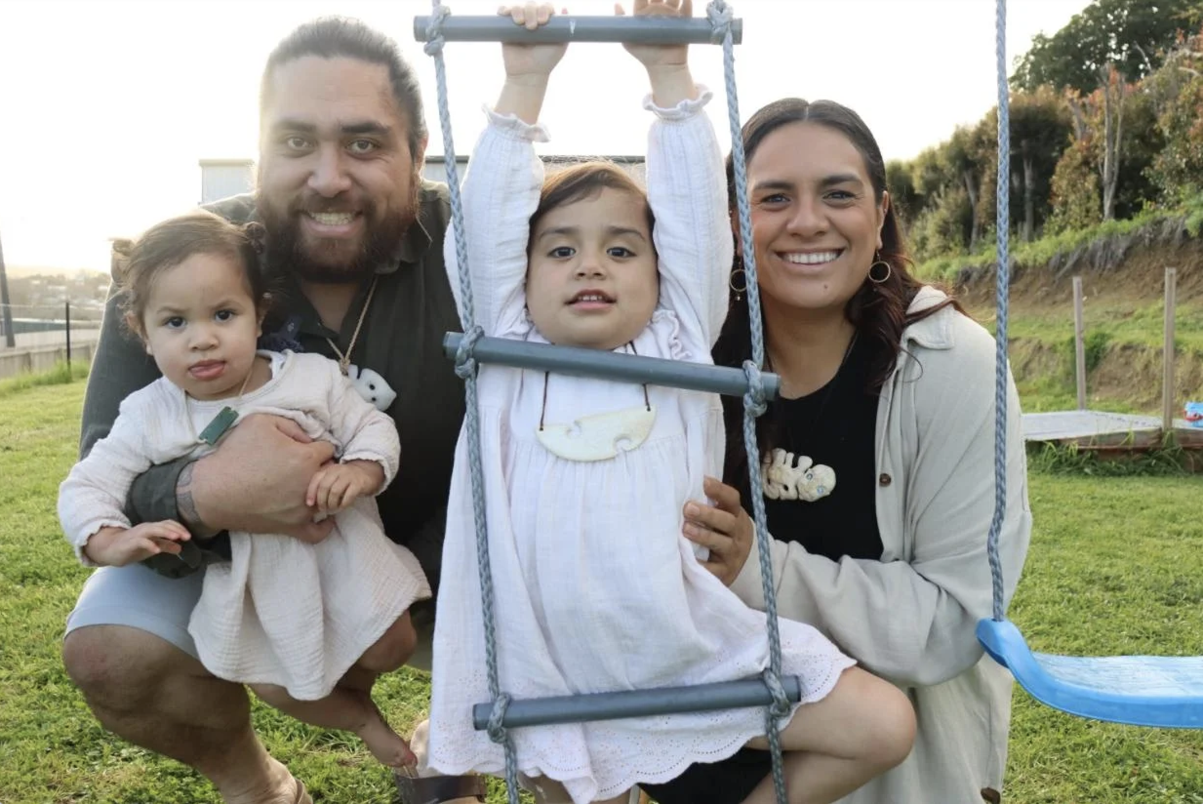 A family of four outdoors on a grassy area, smiling at the camera. A man with a beard and a woman are holding two young girls; one girl is sitting in a blue swing, and the other is in the man's arm. The background shows trees and a slightly hilly landscape.