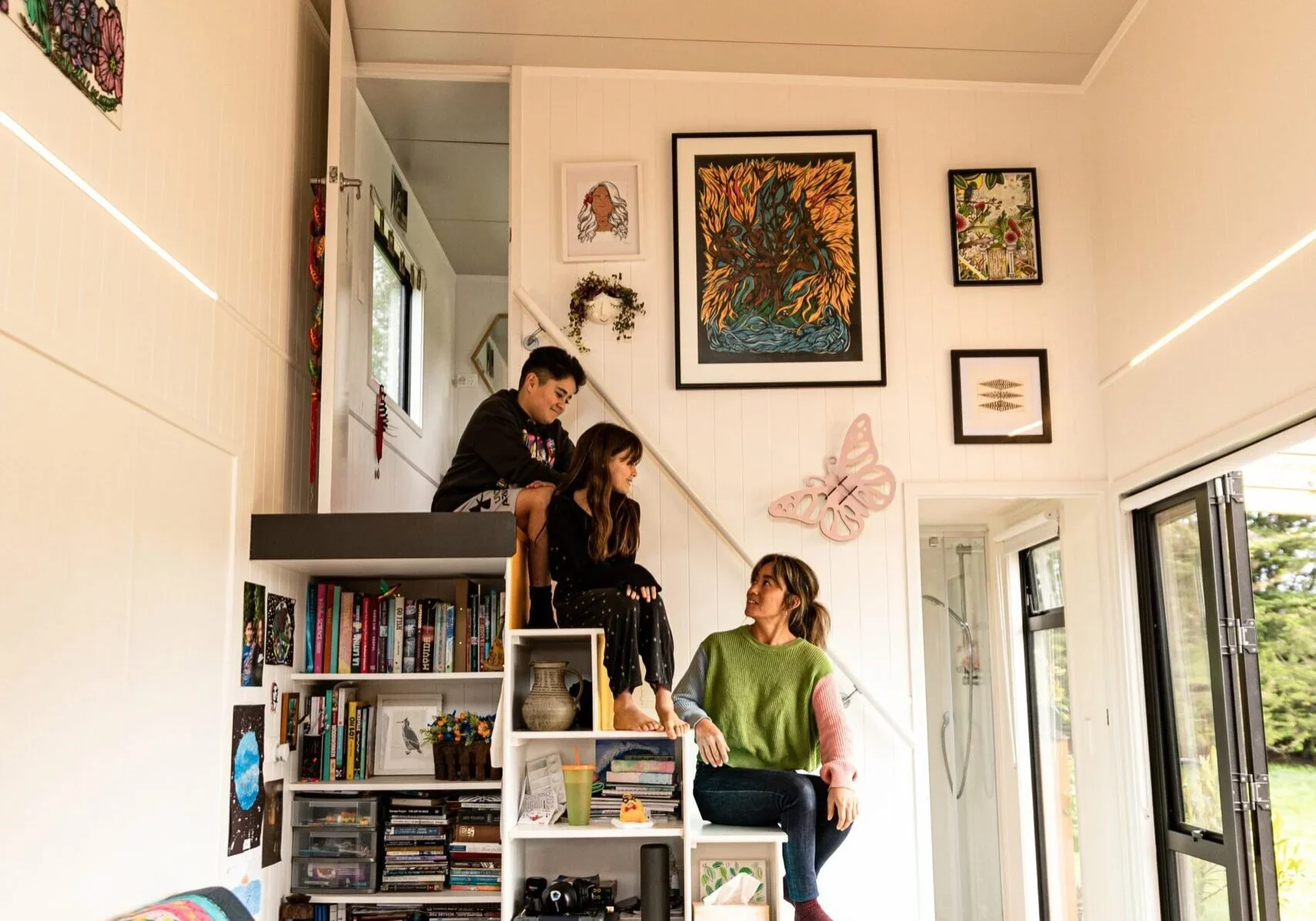 A woman sitting on a white shelf in a bright room, with two children and another child sitting on a higher shelf, near a ladder. The room has white walls with colorful artwork and a butterfly decoration, large windows, and an open door showing outdoor greenery.