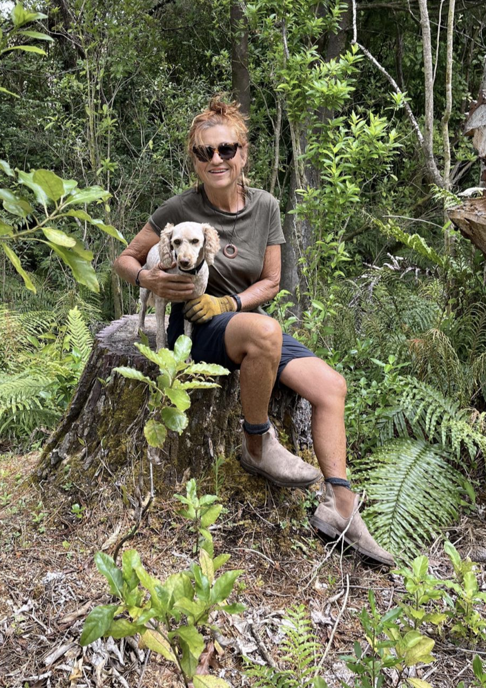 A woman sitting on a tree stump in a forested area, holding a small dog.