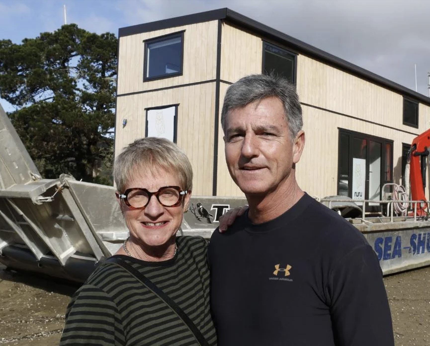 Smiling woman with short blonde hair, glasses, and a striped black and green shirt standing next to a man with short gray hair wearing a black Under Armour shirt, in front of a building under construction or renovation with a boat and trees in the background.
