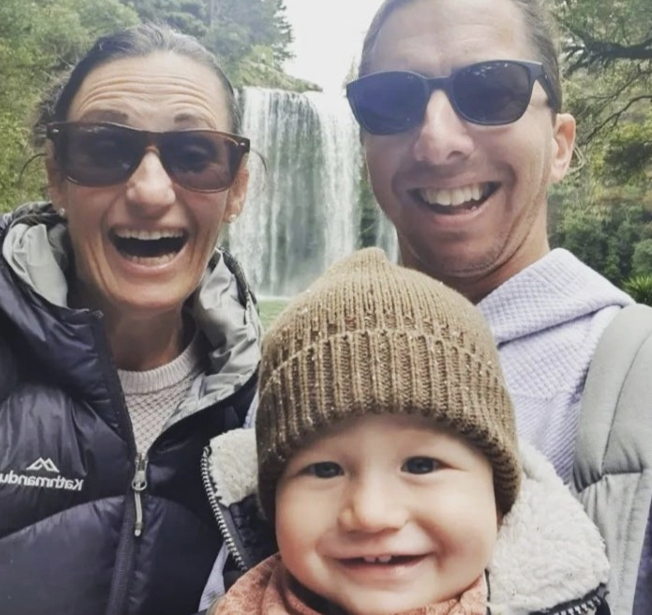 A smiling family of three, including two adults and a young child, taking a selfie outdoors in front of a waterfall surrounded by trees, all wearing casual outdoor clothing and sunglasses.