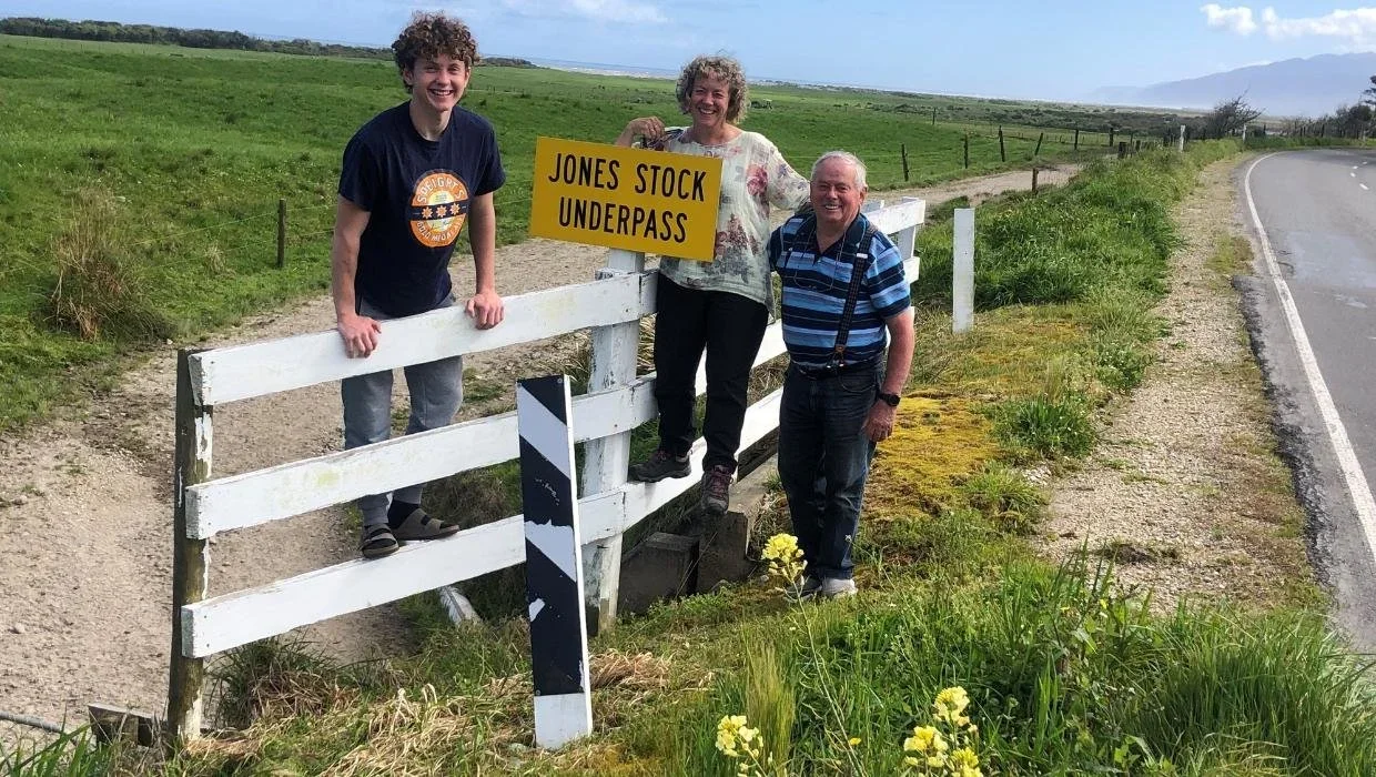 Three people standing near a white fence next to a road in a rural area, holding a yellow sign that reads 'JONES STOCK UNDERPASS' and smiling.
