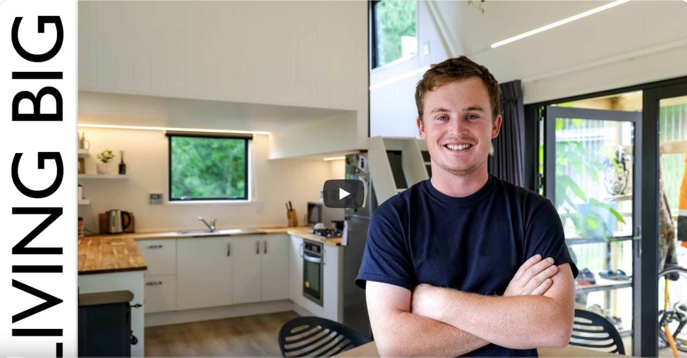 Young man smiling with arms crossed standing in a modern kitchen with large windows and a sliding glass door.