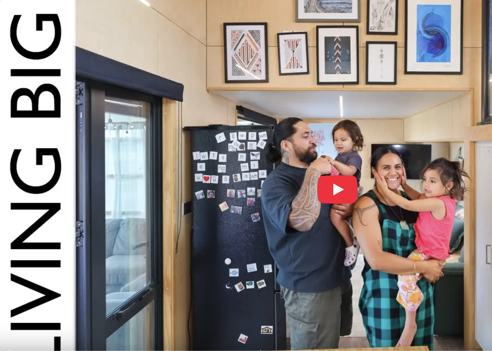 A family of four inside a modern home, with a man holding a young girl, and a woman holding a young girl, all smiling and interacting. The background features framed artwork on the wall, a black refrigerator with magnets and photos, and a window with a curtain.
