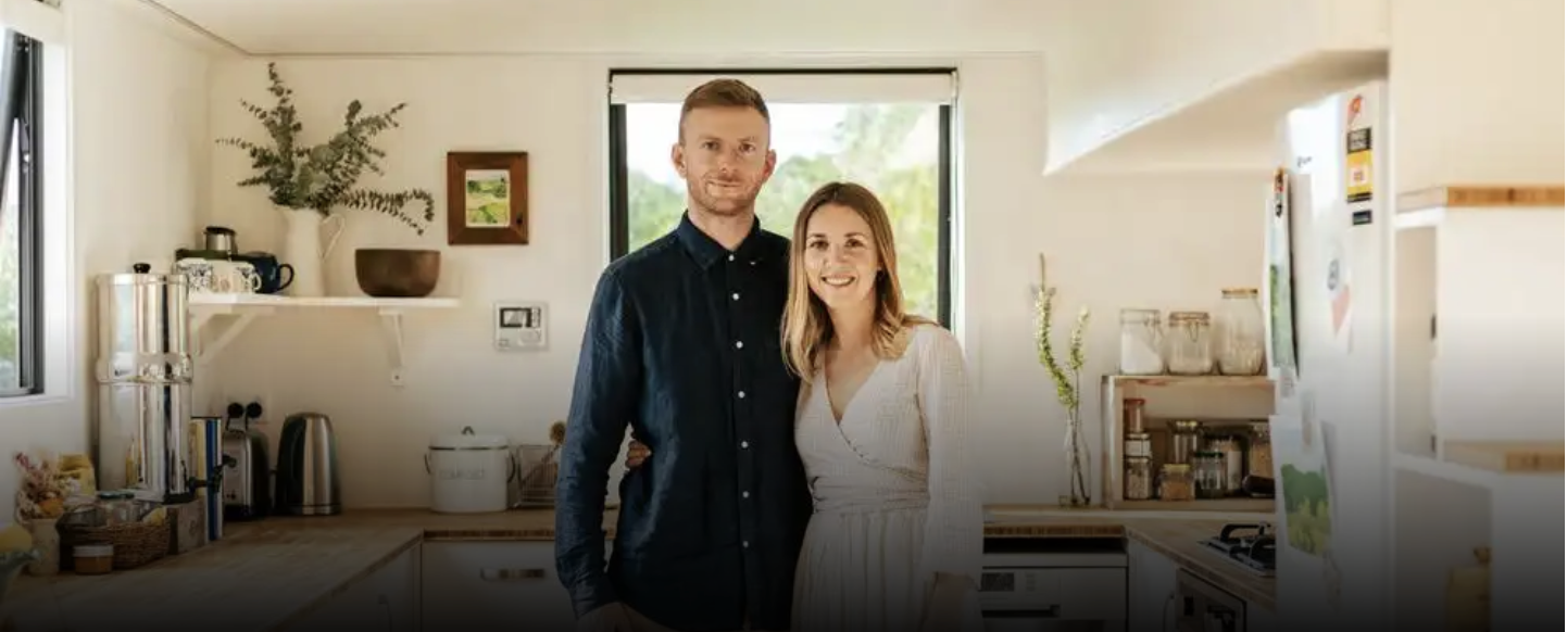 A smiling couple stands in a bright, modern kitchen, with the man wearing a dark blue shirt and the woman wearing a white dress.