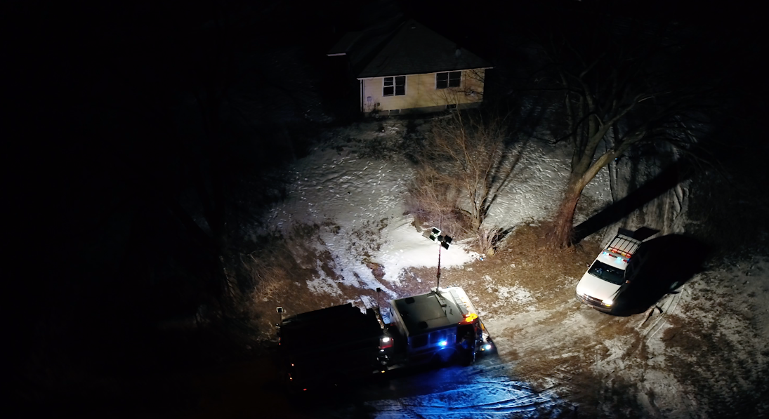 Nighttime scene with police vehicles, floodlights, snow-covered ground, trees, and a house.