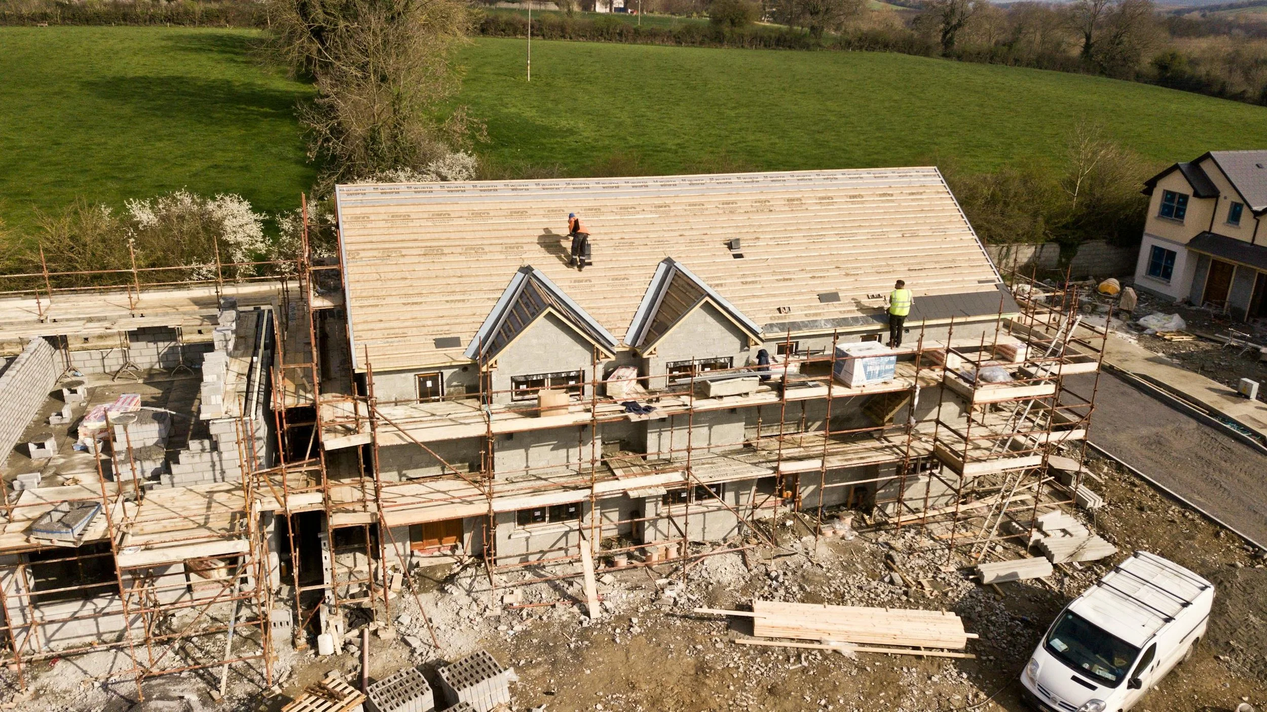 Aerial view of a house under construction with scaffolding around it. The roof is partially built with workers present, and there is a green field in the background.