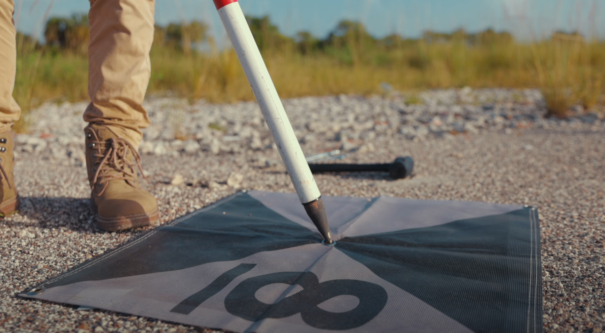 Close-up of a person wearing tan boots and beige pants using a white pole with a pointed tip to mark a spot on a black and gray survey flag with the number 18 written on it, outdoors on gravelly ground.