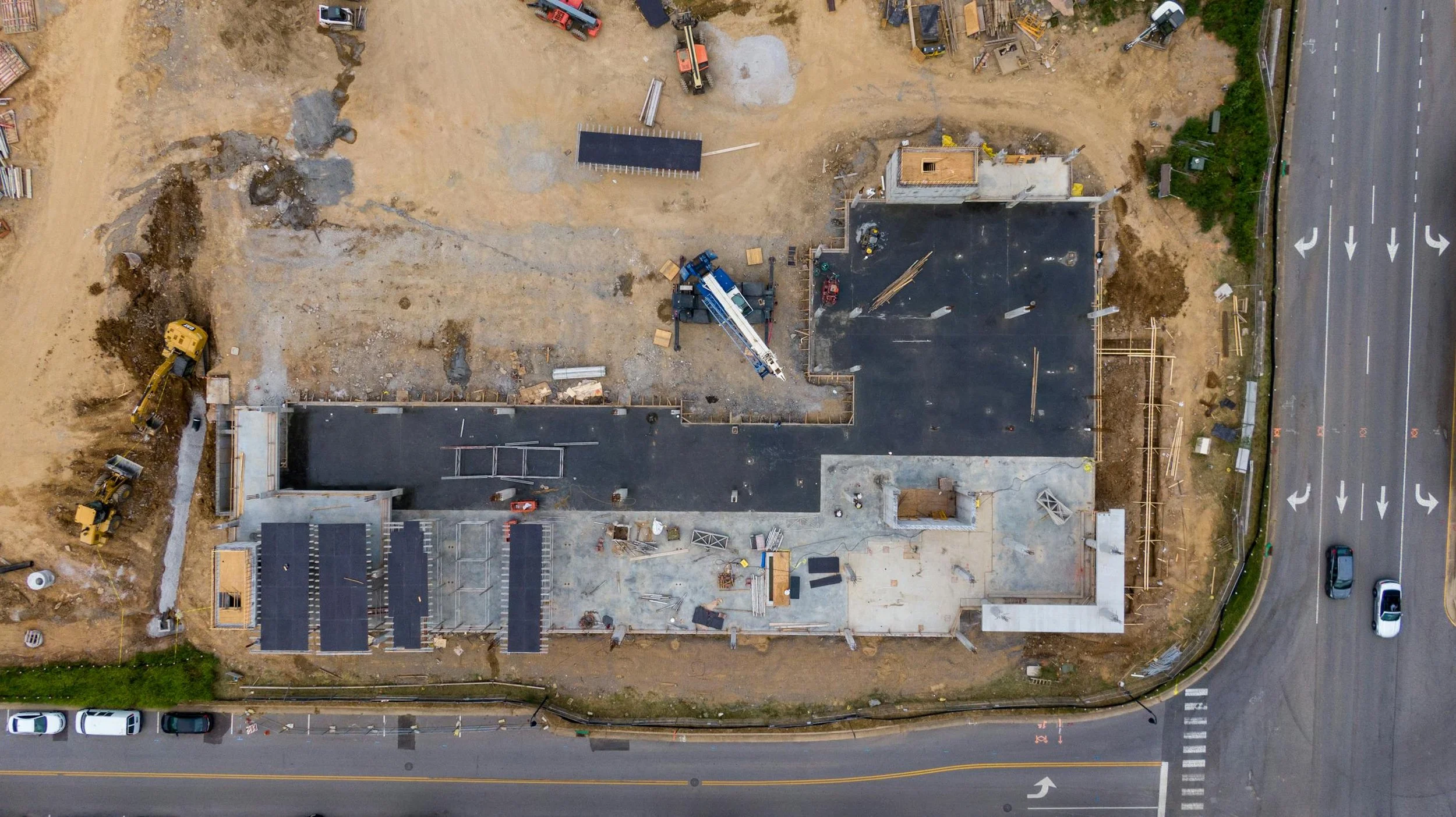 Aerial view of a building construction site with construction vehicles and materials, adjacent to a road with moving and parked cars.