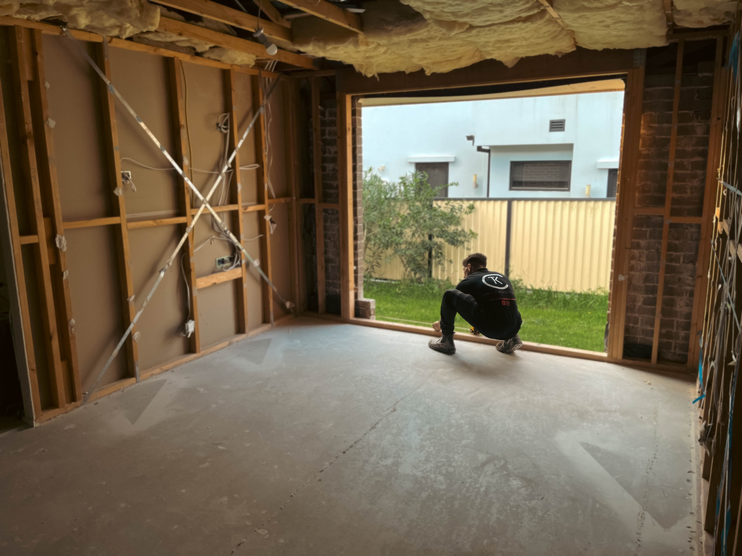 Construction worker kneeling inside a house with unfinished walls, looking out through a large window opening at a yard with grass, trees, and neighboring buildings.