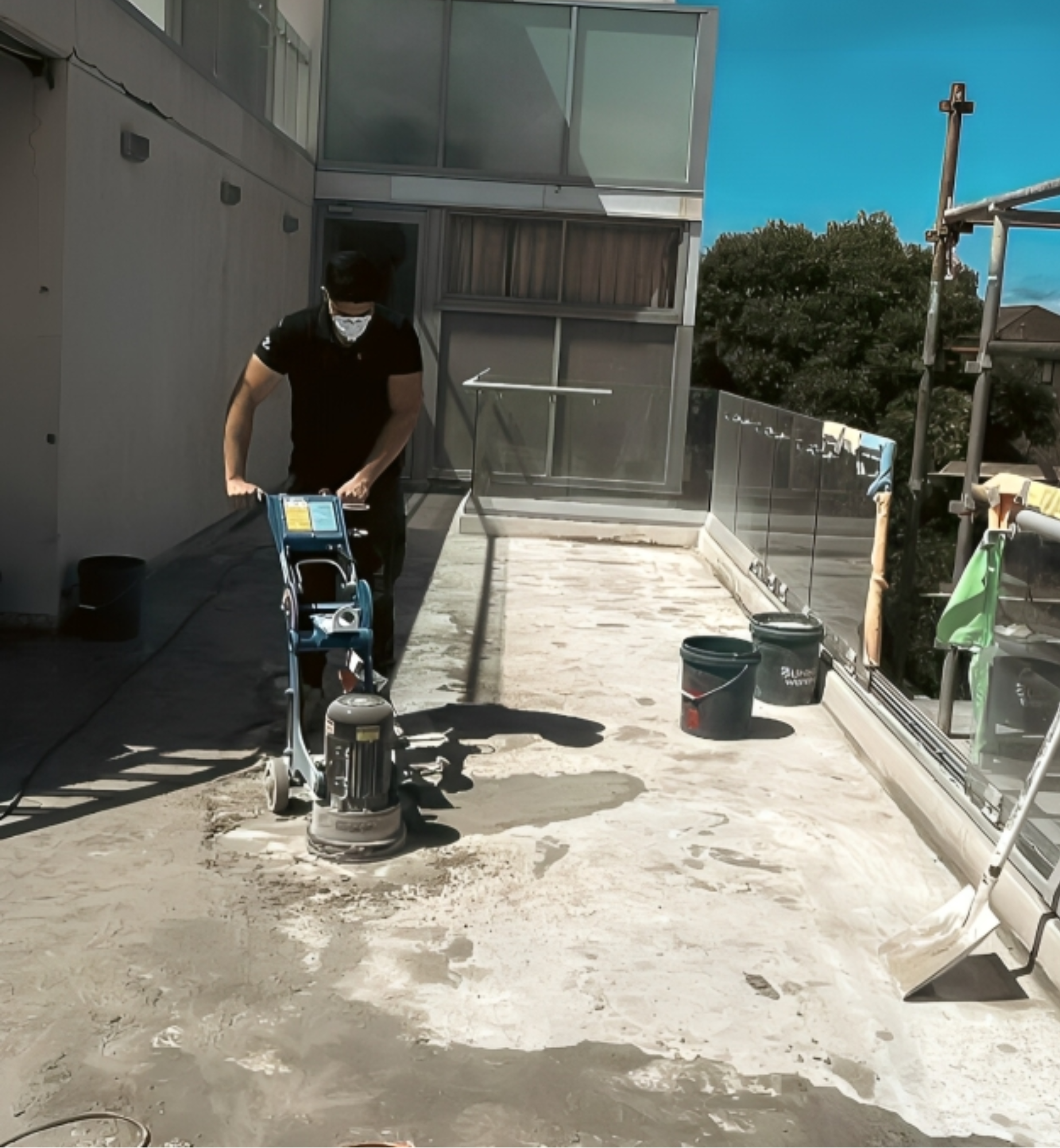 A person wearing a black shirt and face mask operating a floor grinder on a rooftop surface with buckets and construction supplies around.