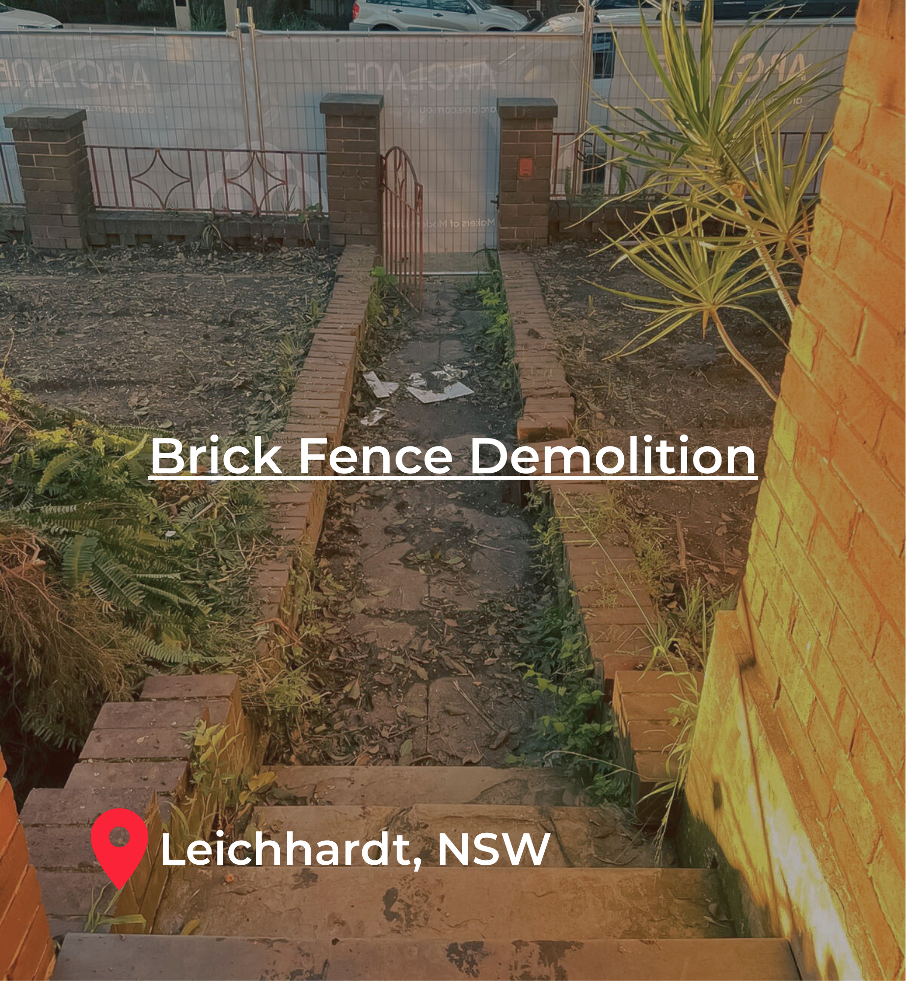 A backyard with a brick fence being demolished, showing dirt and debris on the ground, plants on the sides, stairs leading down, and a street with parked cars in the background in Leichhardt, NSW.