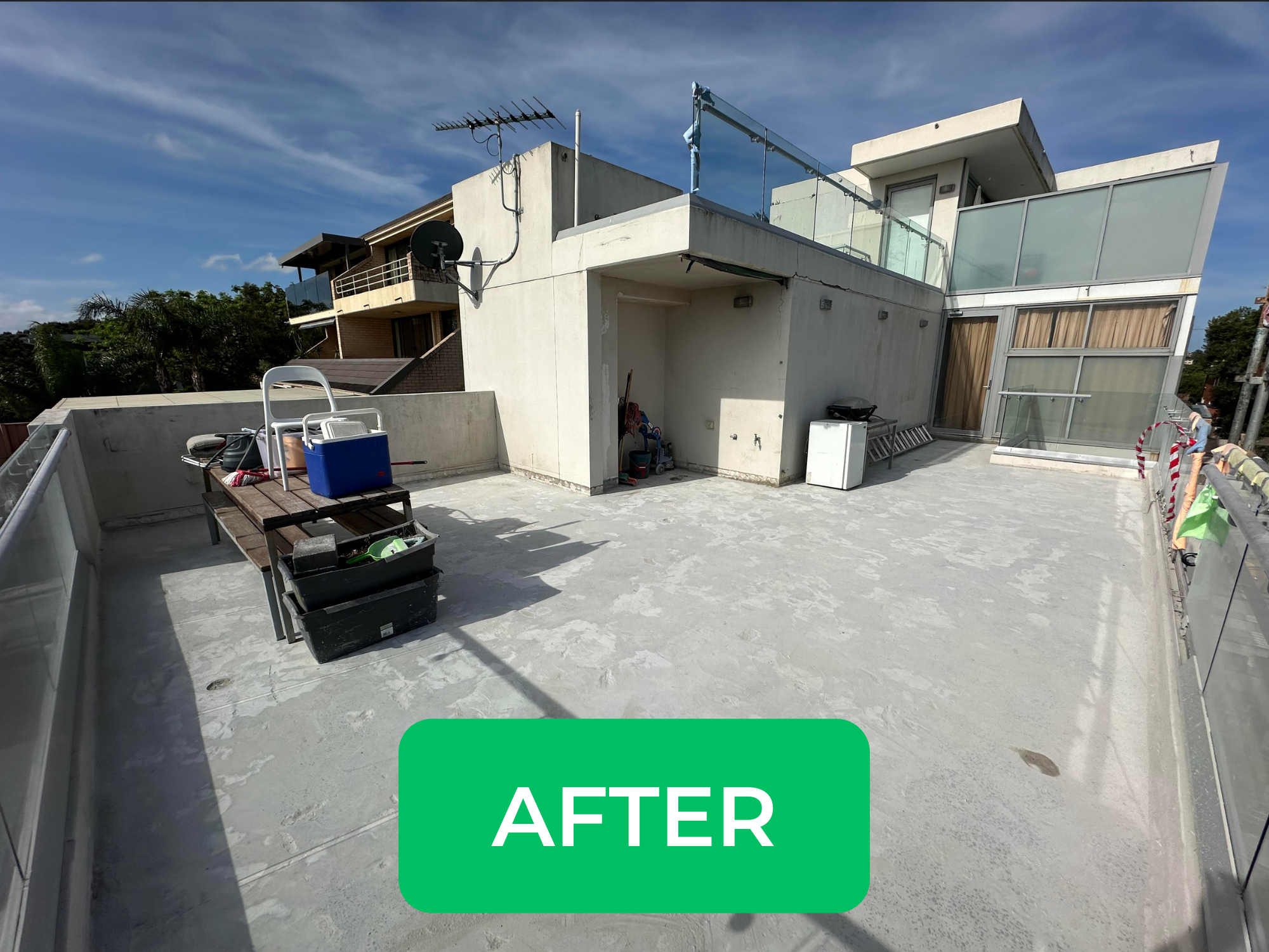 Photo of a clean, recently renovated rooftop terrace with a concrete floor, surrounded by modern glass and concrete railing, and a view of neighboring residential buildings under a partly cloudy sky.