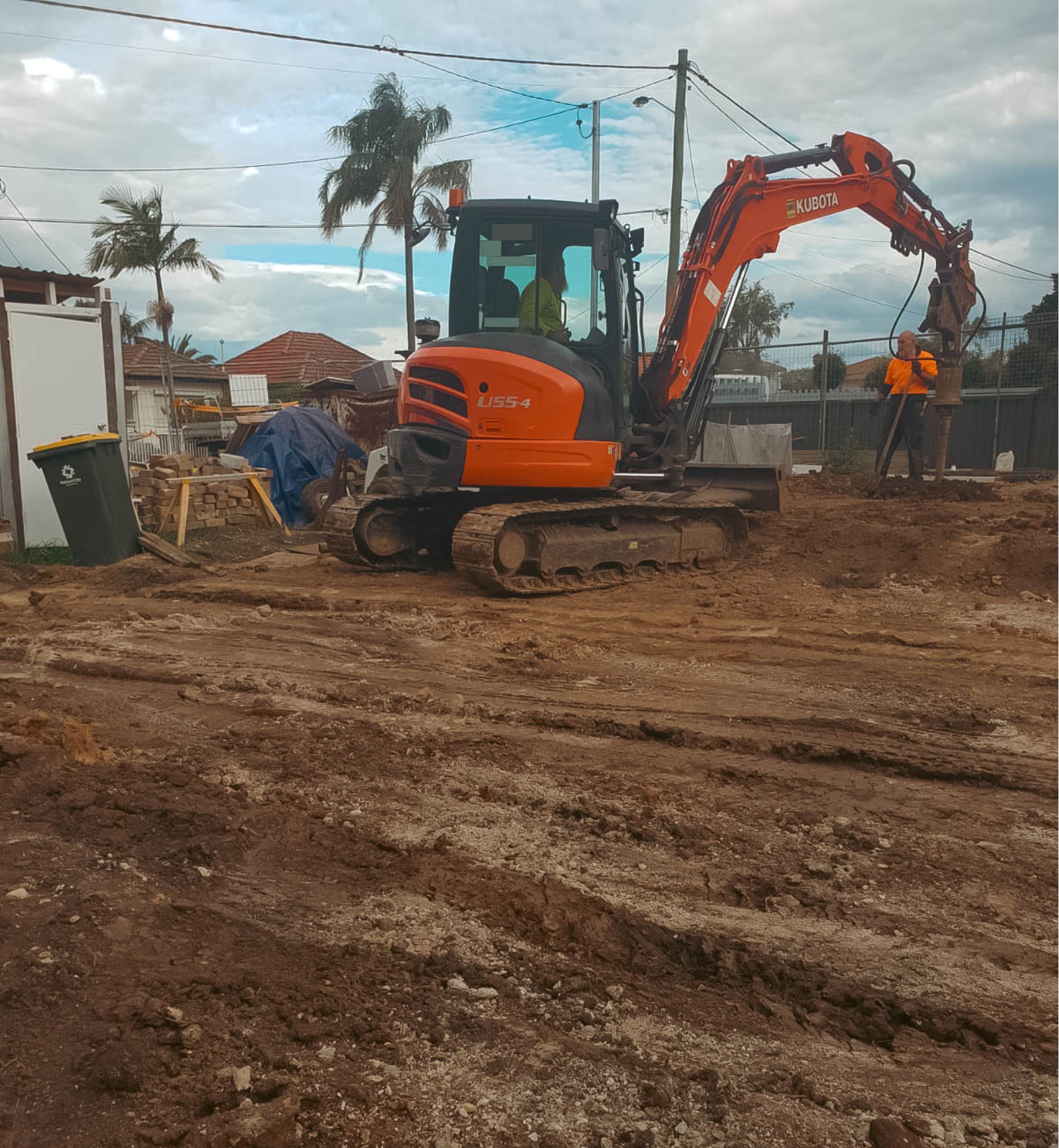 Construction site with a small orange Kubota excavator and two workers, one operating the excavator and the other standing nearby with a shovel. The ground is uneven dirt.
