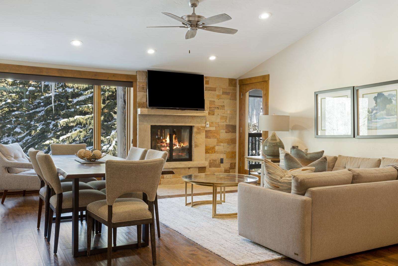 Living room with fireplace, flat-screen TV, beige sofa, armchair, round coffee table, dining table with chairs, large window showing snowy trees, ceiling fan, artwork on wall, and a table lamp.