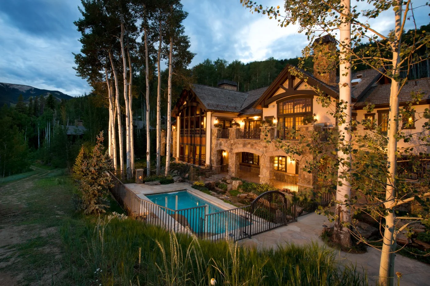 A large house with stone and wooden exterior walls, multiple lit windows, and a balcony with outdoor lighting. There is a small fenced swimming pool in the backyard, surrounded by trees and landscaping, with a mountain and forest in the background during dusk.