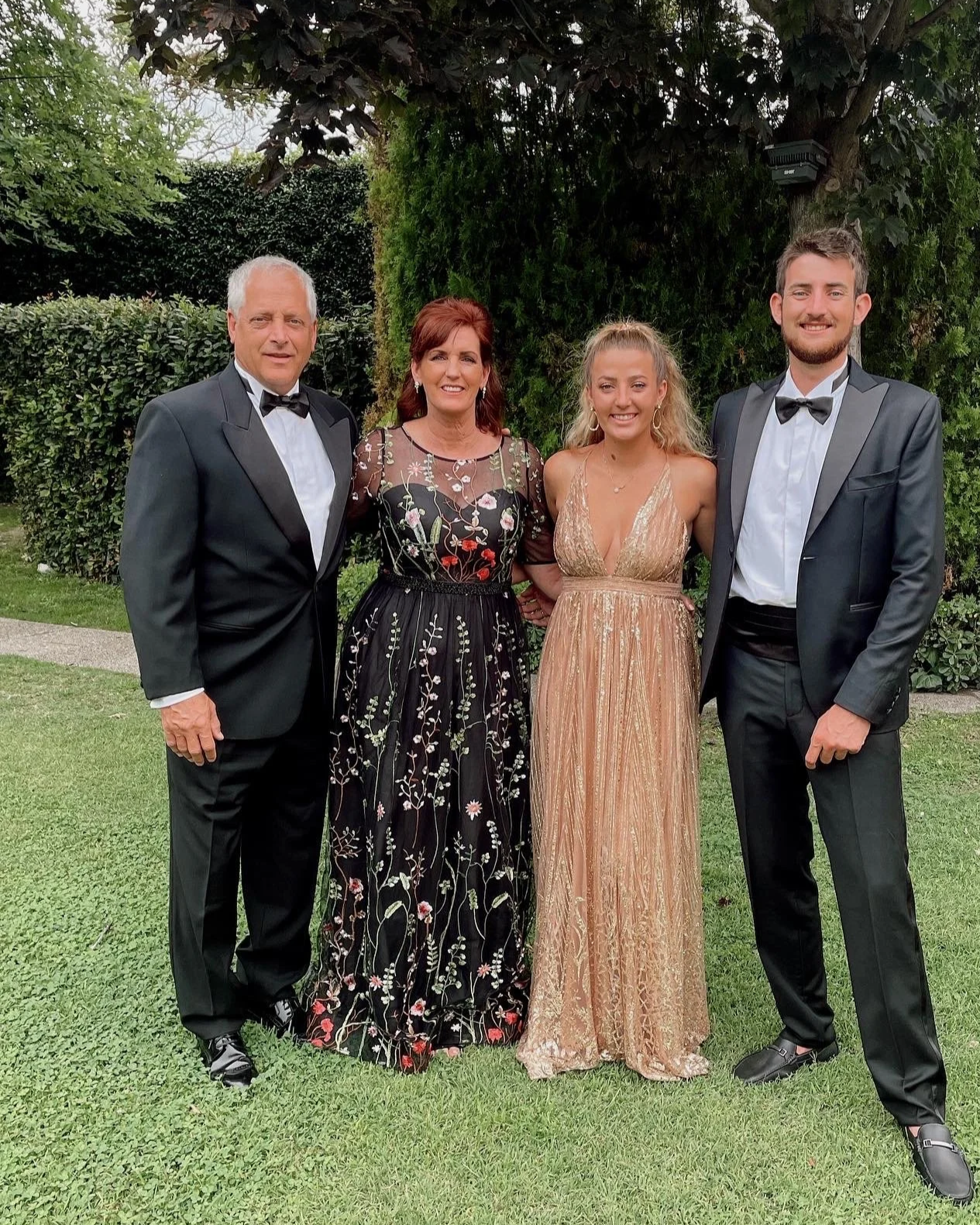 Four people dressed in formal wear standing outdoors in front of green shrubbery, posing for a photo on a grass lawn.