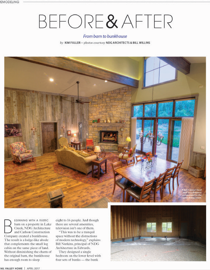 The image shows the interior of a renovated living room with a rustic style. It features wooden walls, a stone fireplace, large windows letting in natural light, a ceiling fan, and wooden furniture, including a dining table and chairs. There is a cozy sitting area with sofas and armchairs, and the room has warm lighting and decorative accents.