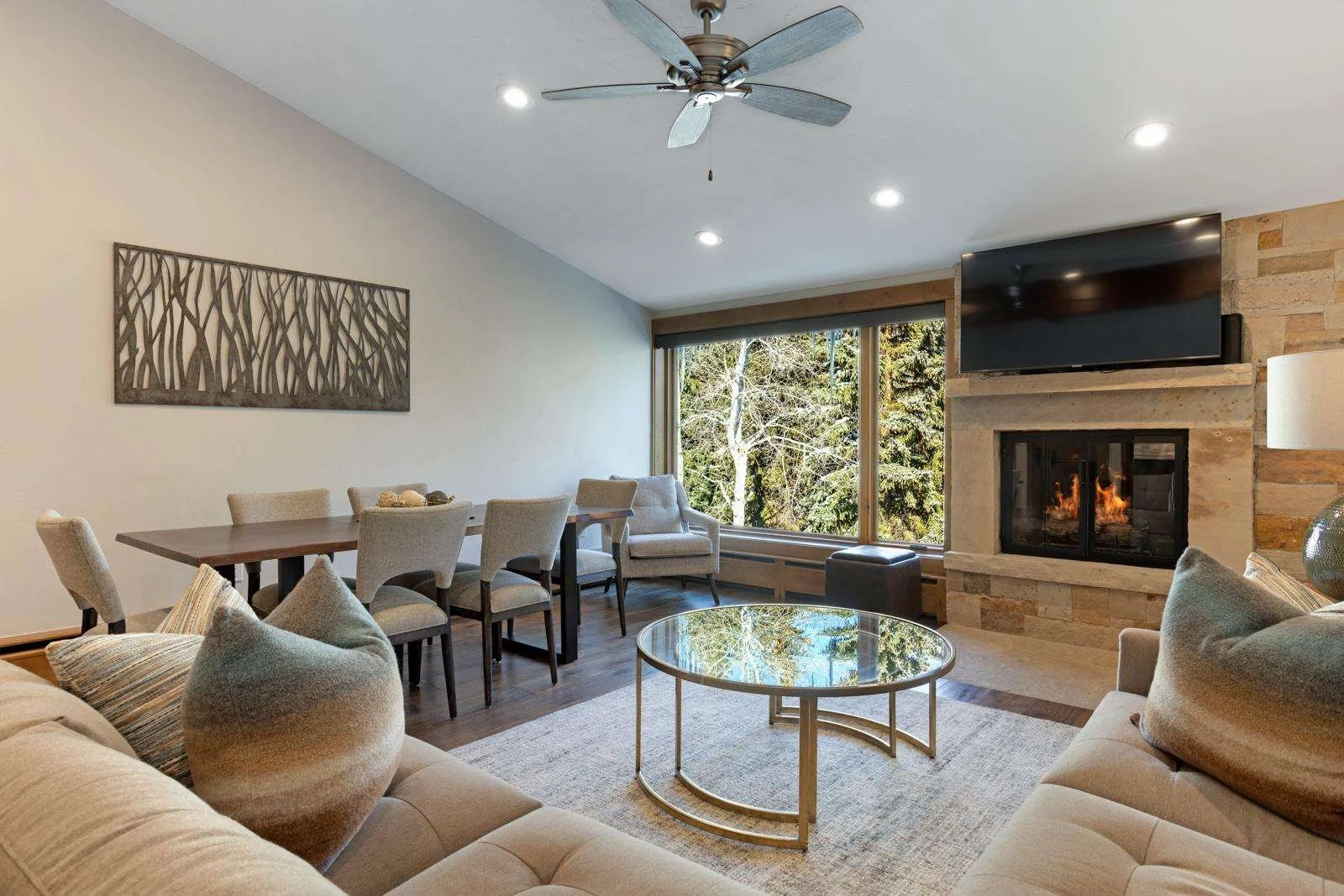 Living room with beige sofa and armchair, round glass coffee table, stone fireplace with TV mounted above, large window showing trees outside, wood floor, ceiling fan, abstract wall art, and a light-colored area rug.