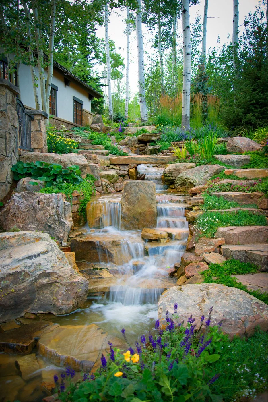 A cascading waterfall flowing over rocks surrounded by lush green plants and colorful flowers, with a house and tall trees in the background.