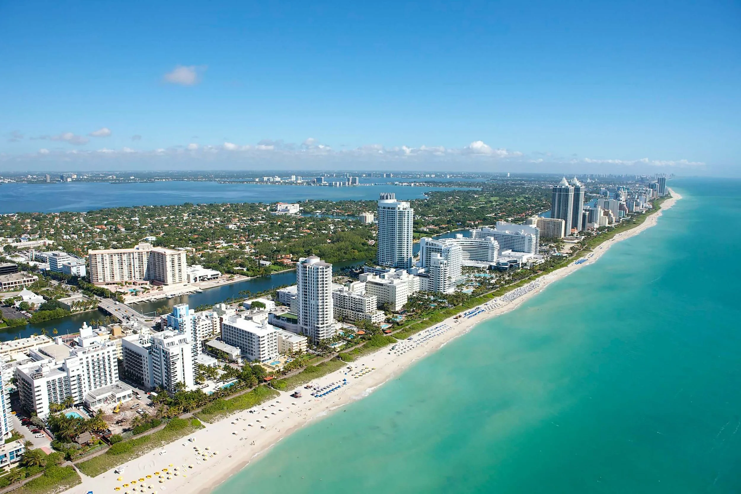 Aerial view of a coastal city with high-rise buildings along a white sandy beach, turquoise ocean, and a bay with calm water and boats, under a blue sky with some clouds.