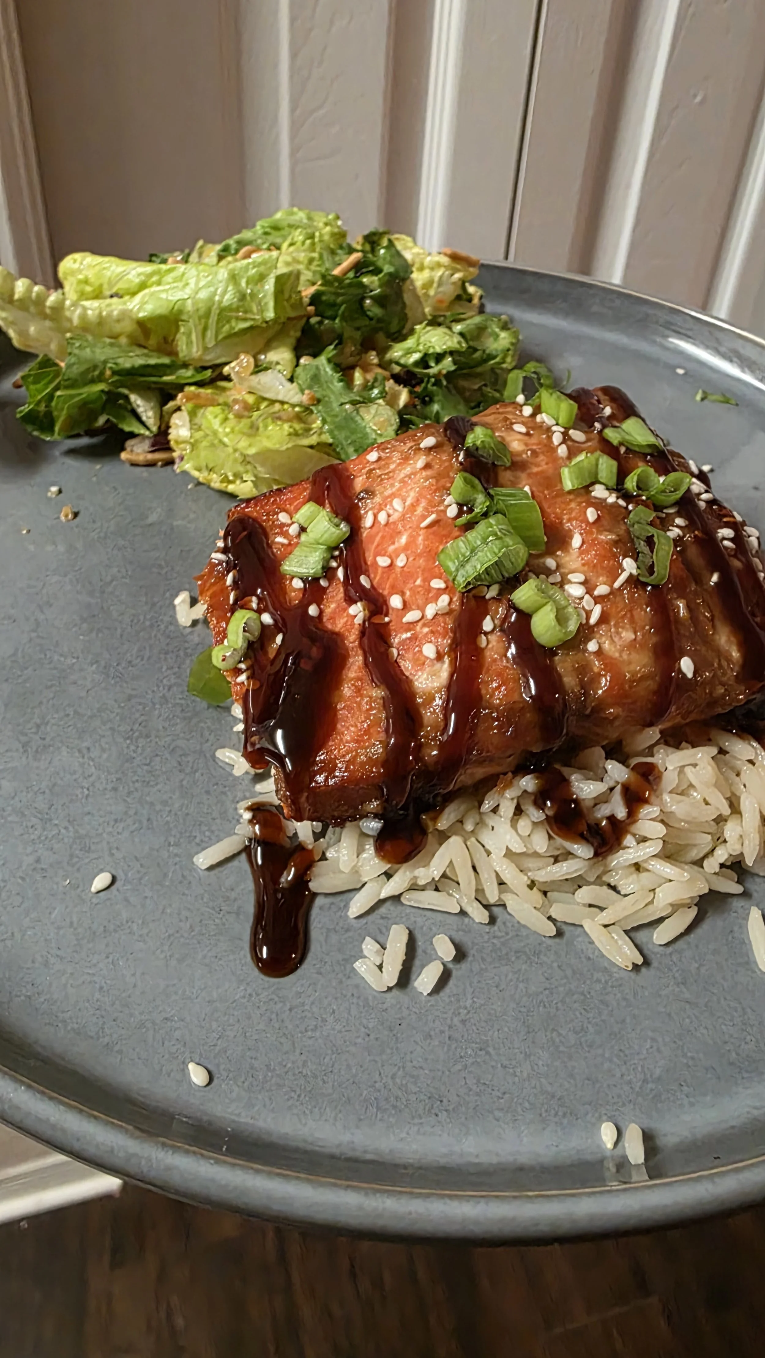 A plate with rice topped with a glazed piece of salmon, garnished with green onions and sesame seeds, with a side of chopped green salad.