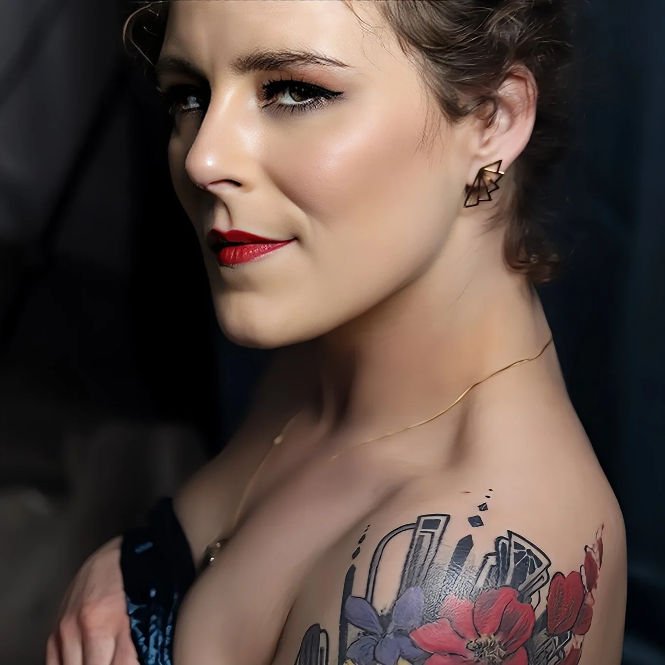 A close-up of a woman with short hair, red lipstick, and makeup, wearing geometric earrings and a gold necklace. She has a tattoo of red and purple flowers on her shoulder.