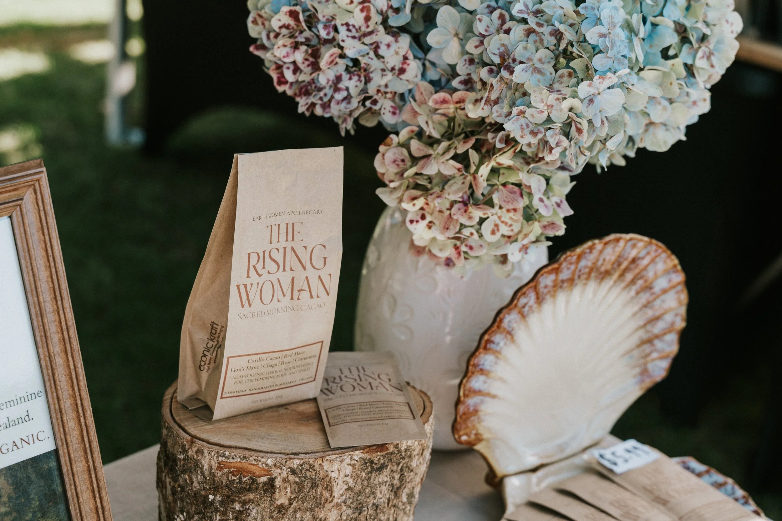 Close-up of a bouquet of pink and white hydrangea flowers in a white vase, with a brown paper bag next to it that reads, "The Rising Woman Sacred Morning Cacao." There is a wooden slice underneath the paper bag and part of a decorative shell is visible on the right. A framed item is on the left side of the image.