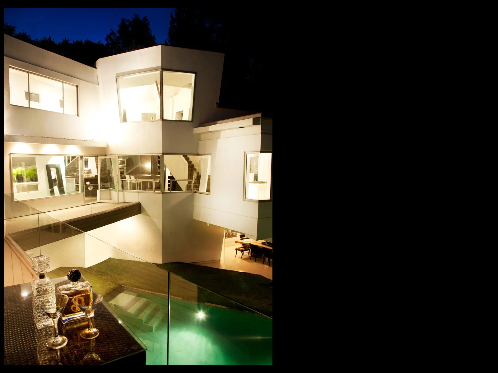 Presented by Missig Team, a real estate team at Berkshire Hathaway HomeServices California Properties in Beverly Hills and Los Angeles. Night view of a modern multi-story house with large illuminated windows, showcasing an interior staircase and livi