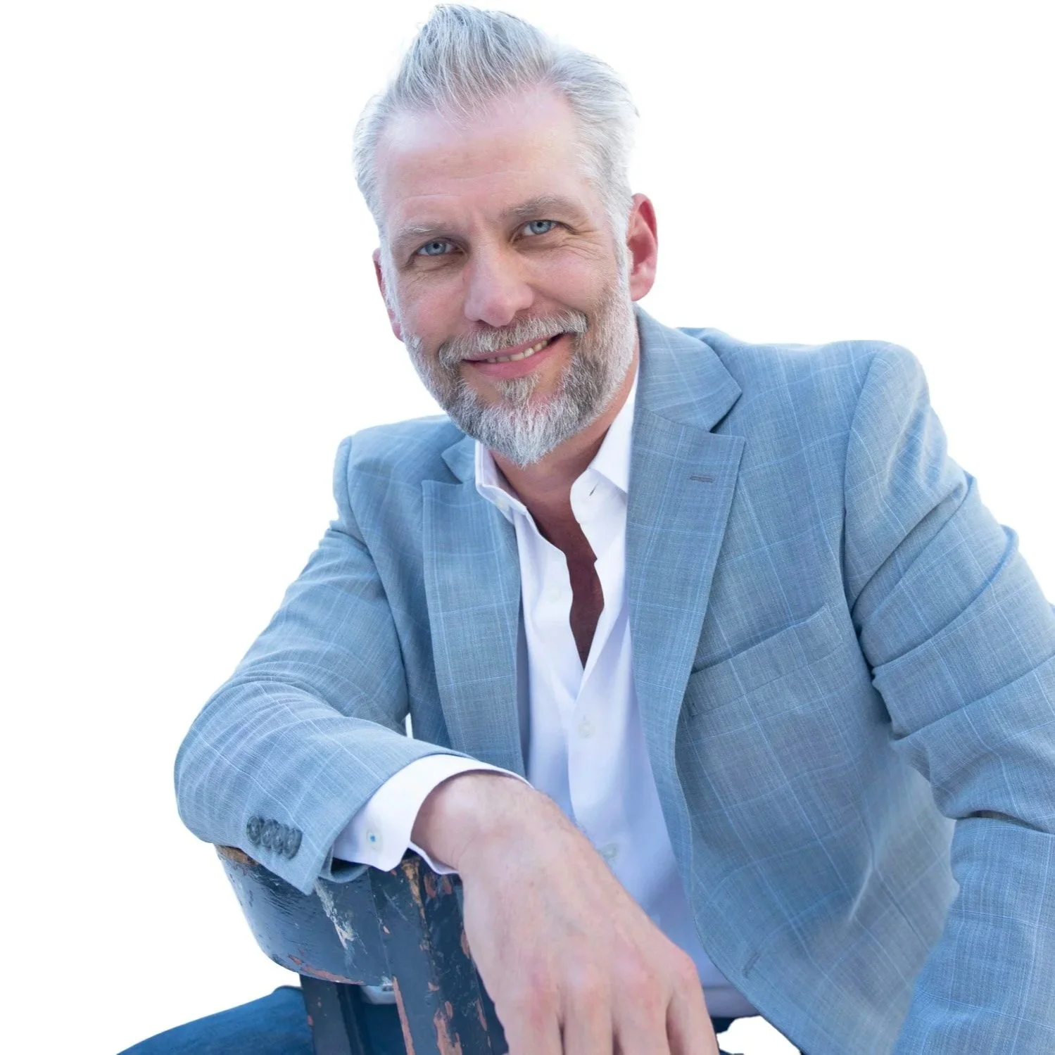 A smiling Phil Missig, wearing a light blue blazer and white shirt, leaning on a chair against a white background.