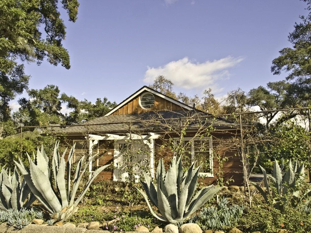 Presented by Missig Team, a real estate team at Berkshire Hathaway HomeServices California Properties in Beverly Hills and Los Angeles. A house with a gabled roof is surrounded by a garden with large succulents and various plants, under a clear blue
