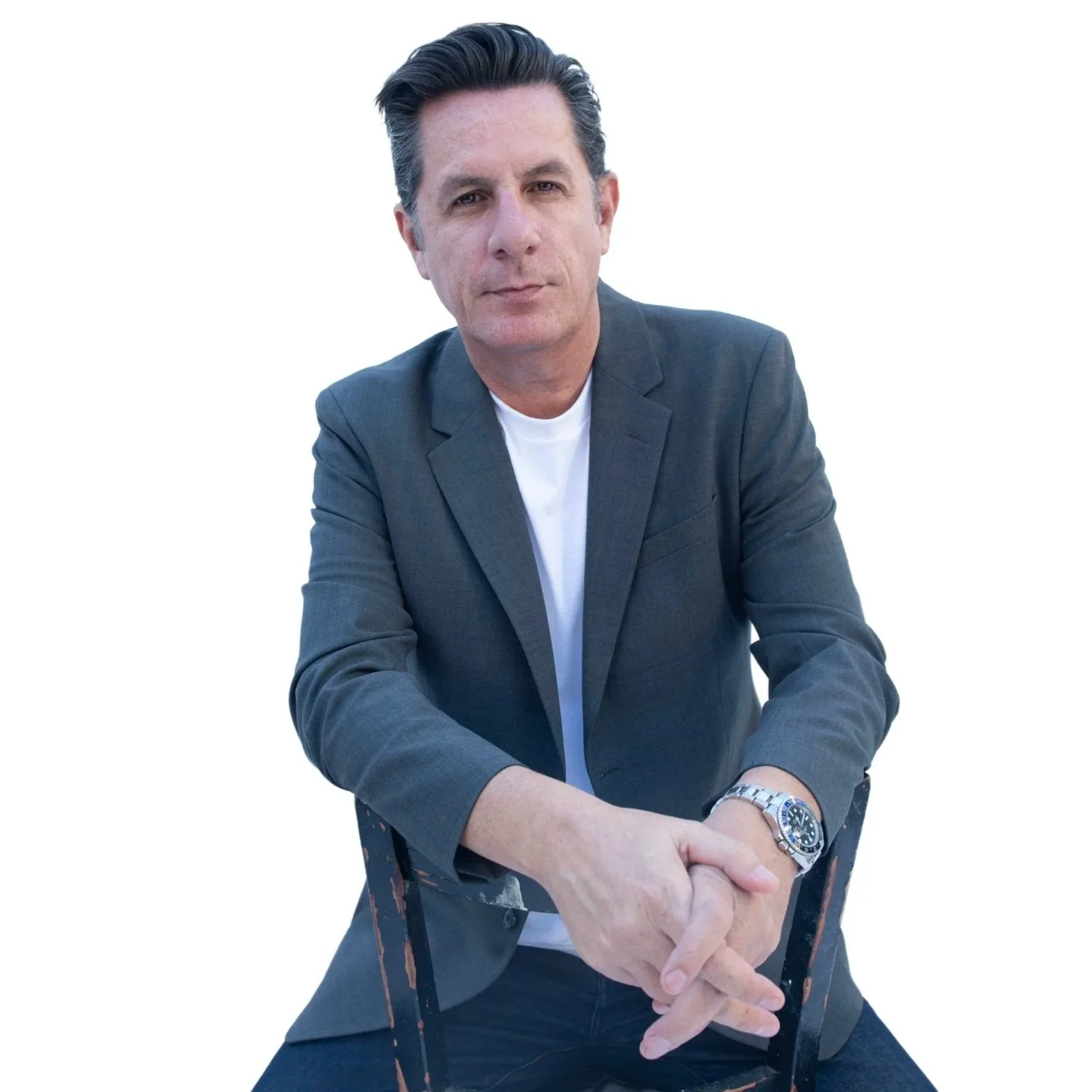 A man in a dark gray blazer and white shirt sitting on a black chair against a white background, with his hands clasped in front of him, wearing a silver watch.