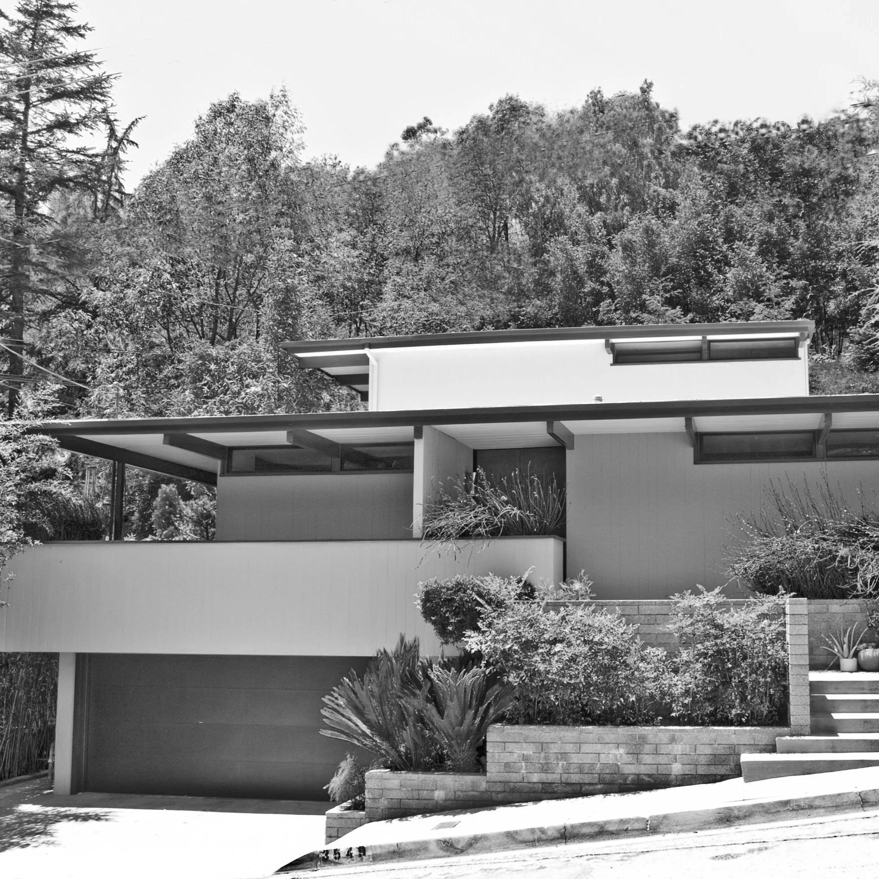 Black and white photo of a modern house with geometric design, flat roofs, and large windows. The house is set on a slope with stairs and surrounded by trees.
