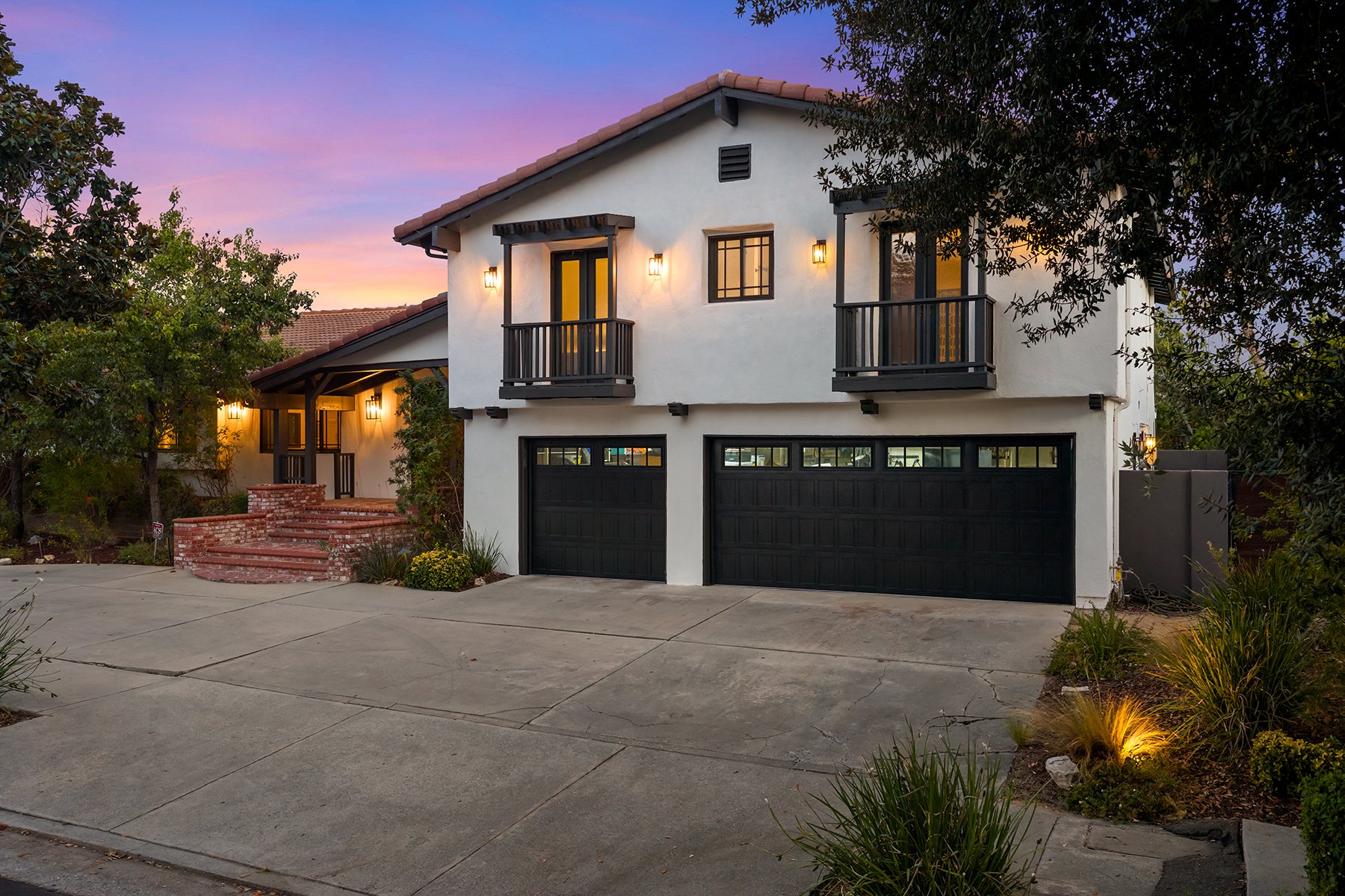 A two-story house with white stucco walls, black window frames, and black garage doors. The house has small balconies with black railings and warm outdoor lighting. There is a brick staircase leading to the front door, surrounded by trees and landscaping, with a sunset sky in the background.