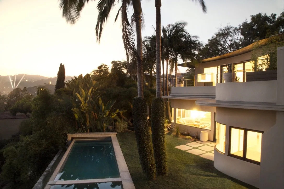 Presented by Missig Team, a real estate team at Berkshire Hathaway HomeServices California Properties in Beverly Hills and Los Angeles. Luxury Streamlined Moderne home with large windows, an infinite swimming pool, palm trees, and lush greenery at su