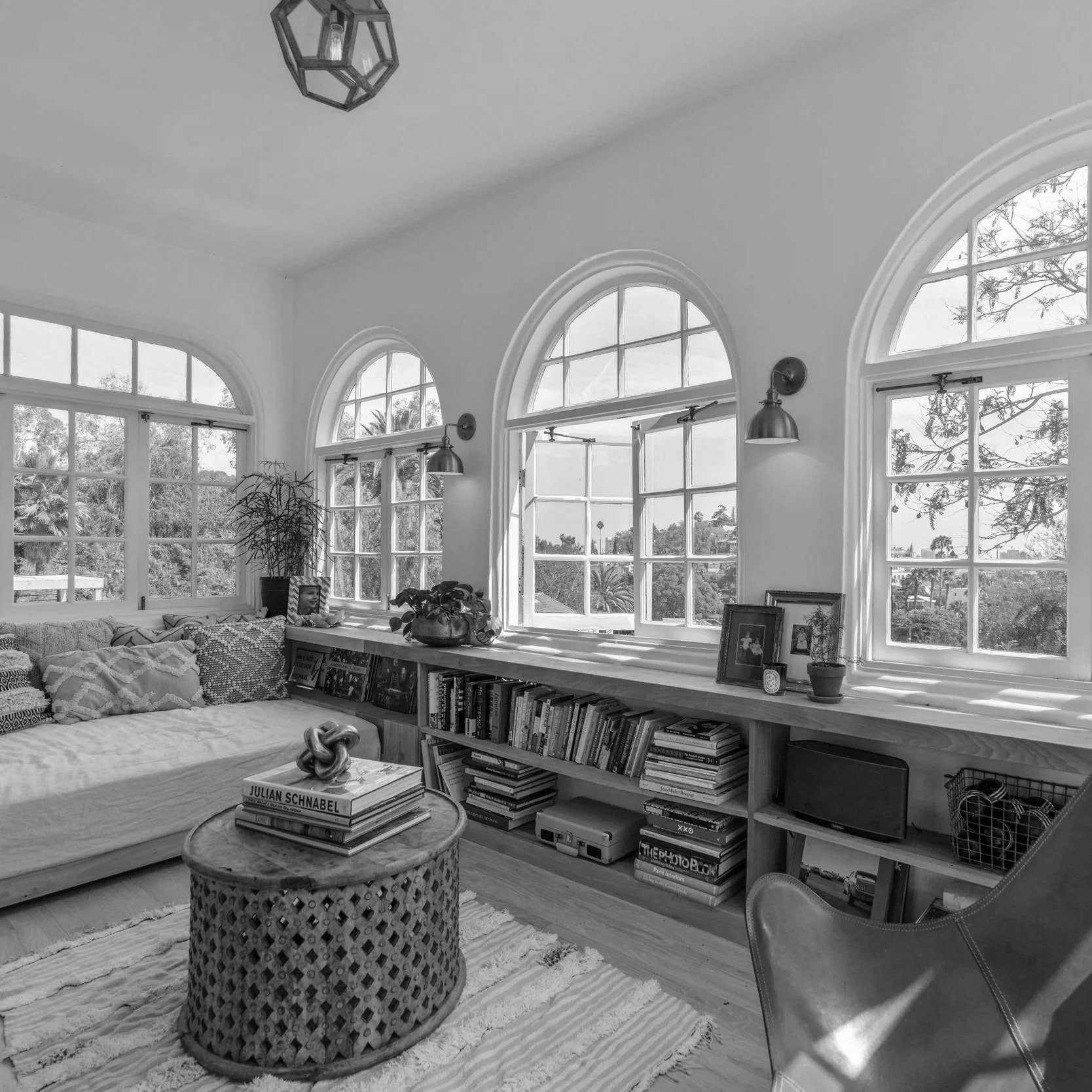 A black and white photo of a cozy living room with large arched windows, a sectional sofa with patterned cushions, a round woven coffee table with books, a built-in bookshelf with books and decorative items, potted plants, and framed photographs. There is natural light coming through the windows, illuminating the room.