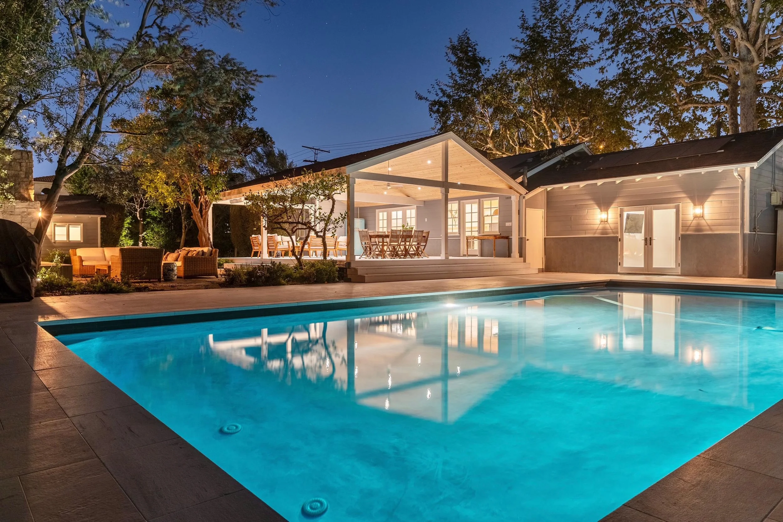 Presented by Missig Team, a real estate team at Berkshire Hathaway HomeServices California Properties in Beverly Hills and Los Angeles. A beautiful backyard pool at dusk with an illuminated house and covered outdoor living room.