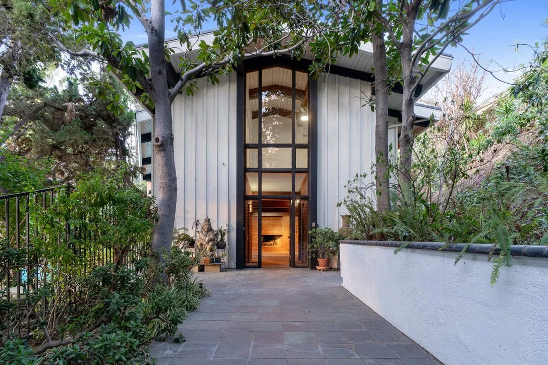 Presented by Missig Team, a real estate team at Berkshire Hathaway HomeServices California Properties in Beverly Hills and Los Angeles. View of a modern post & beam architectural house entrance with tall windows, surrounded by trees and plants, and a